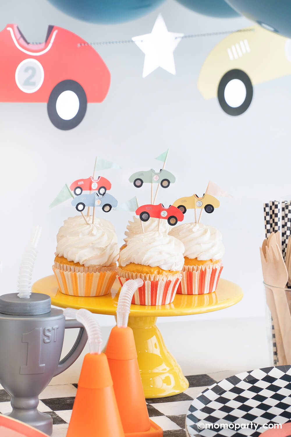 A festive kid's race car party set up featuring some cupcakes decorated with Momopartys's race car cupcake kit that comes with vintage race car toppers and flag toppers in pastel colors, along with orange and red striped cupcake wrappers. Around the cupcakes are some cone shaped and trophy shaped tumbler sippers and round checkered plates, table runner and party straws. Above the table is Meri Meri's race car party garland with some party balloons.