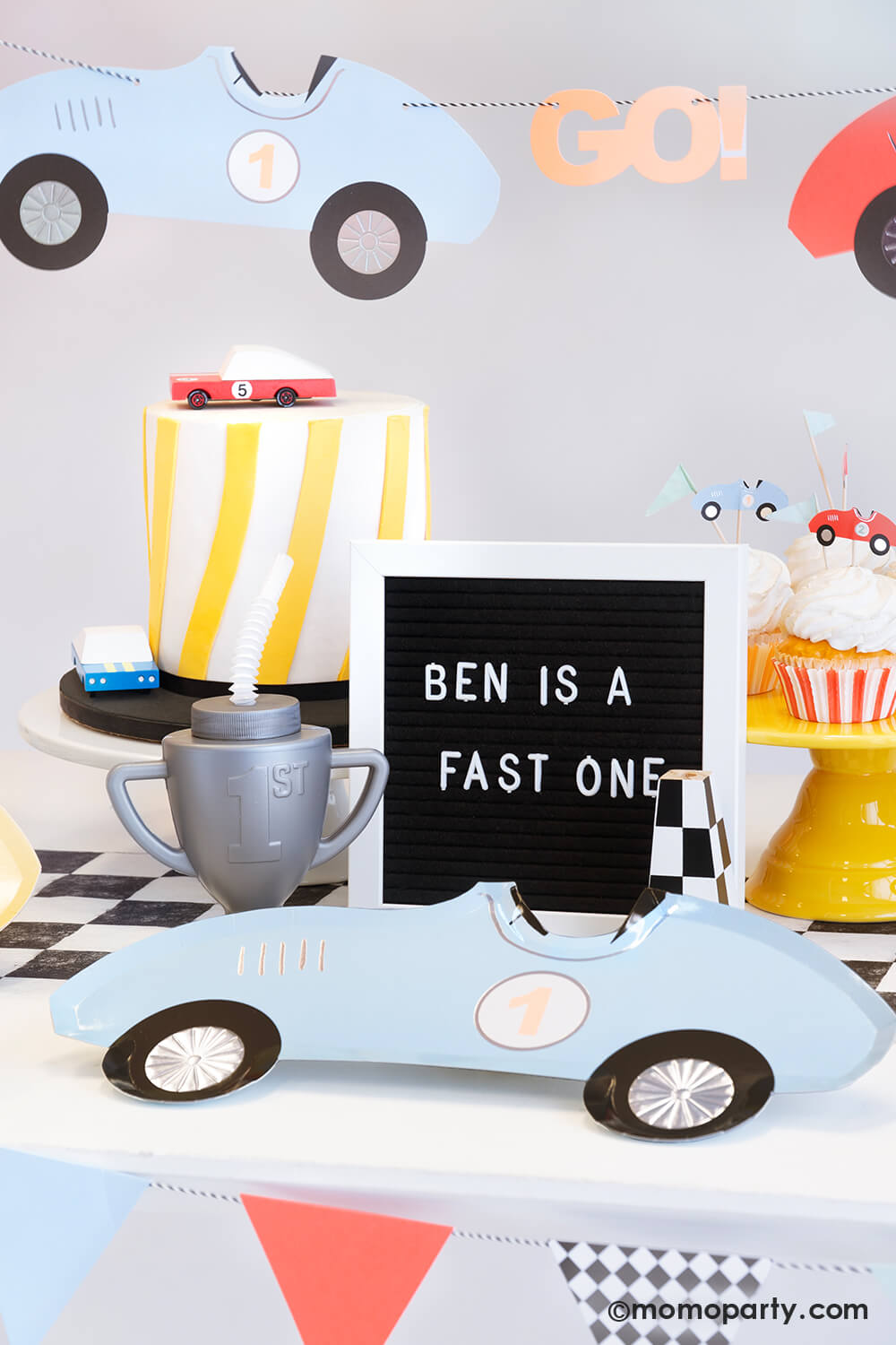 A "Fast One" race car themed first birthday party table featuring Momopartys's race car shaped paper plates, a trophy shaped sipper on a checkered paper table runner, along with a modern cake adorned with wooden toy race cars and some cupcakes decorated with Meri Meri's race car toppers. Above the table hung a vintage race car party garland in soft pastel colors. Along with a letter board spelling "Ben is a fast one" , it makes a perfect inspiration for a baby boy's first birthday celebration.
