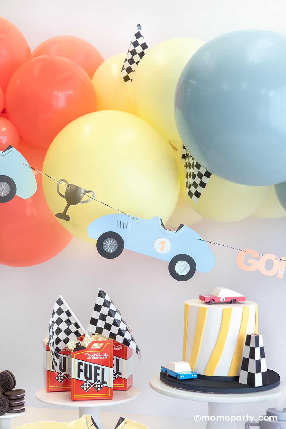 A festive race car themed party set up by Momopartys featuring a pastel balloon garland adorned with checkered flags, underneath hung a vintage race car party garland. On the table are some fuel treat boxes topped with checkered flags and a modern birthday cake topped with a race car wooden toy car toppers - a perfect inspiration for a kid's "Two Fast" second birthday party or "Fast One" first birthday party.