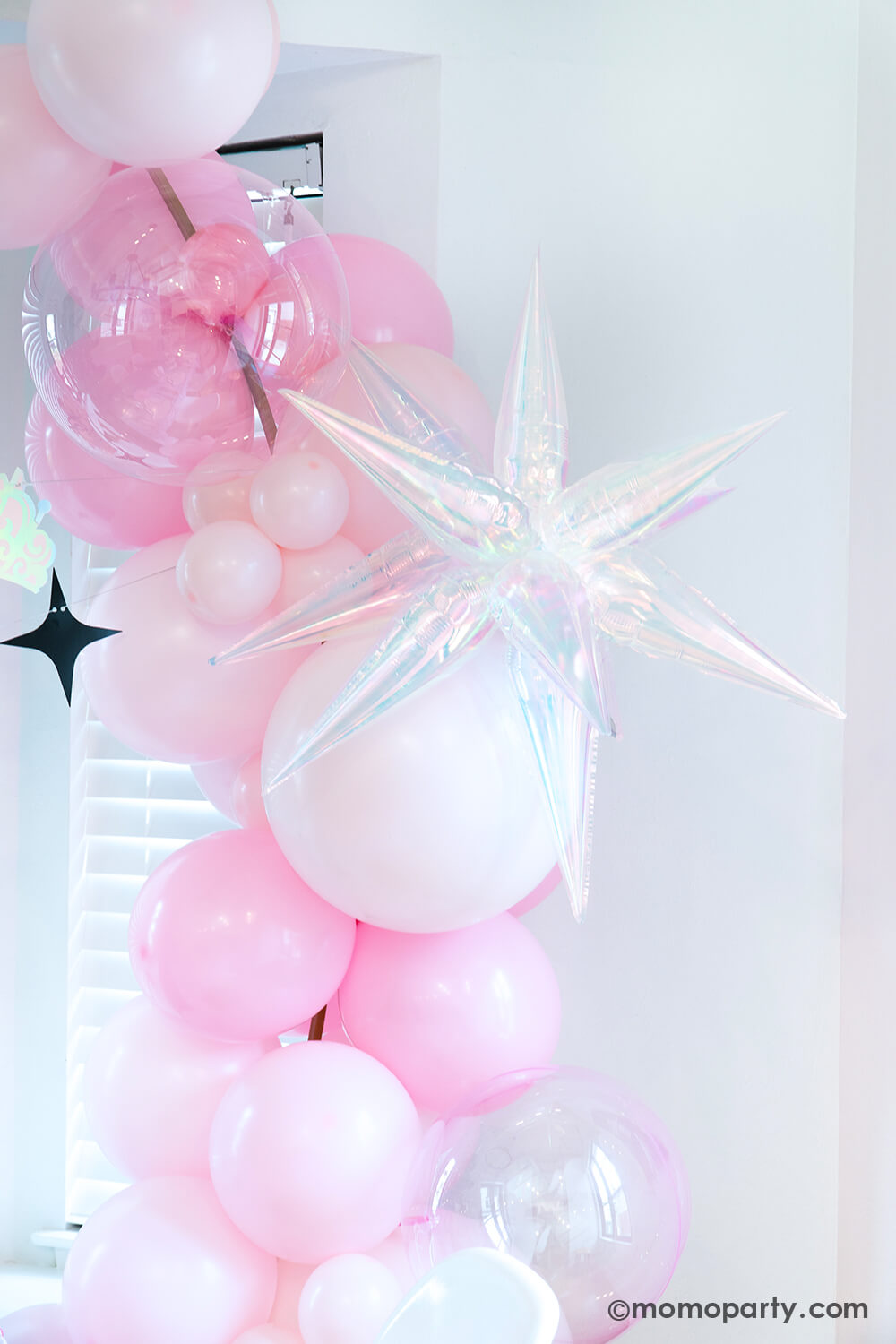 A Wicked Glinda inspired balloon arch by Momopartys with pink and pastel pink latex balloons in various sizes adorned with the 26" iridescent starburst shaped foil balloon and two clear pink orbz non-foil balloons as bubbles next to a window against a white background at a Wicked themed party.