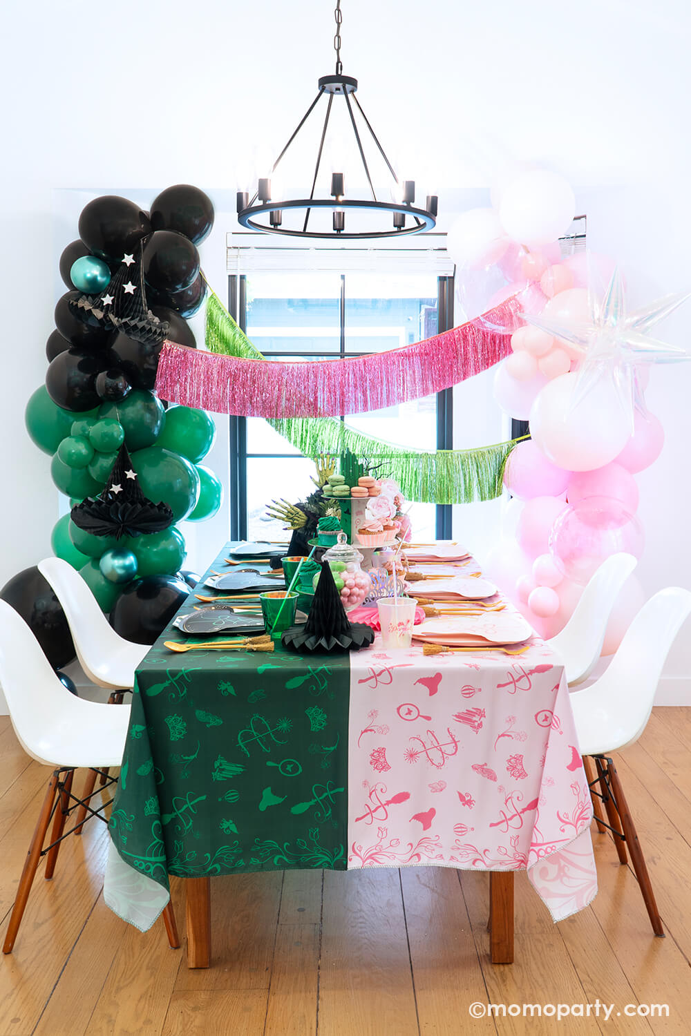 A spellbinding Wicked party set up by Momopartys featuring various Wicked themed party supplies and decorations that represent two worlds or Glinda and Elphaba including pink and black witch hat shaped plates, two-toned Emerald City cupcake stand, Wicked party cups, napkins, witch broom utensil set and Wicked witch hat honeycombs on the green and pink two-tone tablecloth. With Wicked floral arrangement and treats on the table, balloon garlands and pink and green fringe garlands hung above as the backdrop.