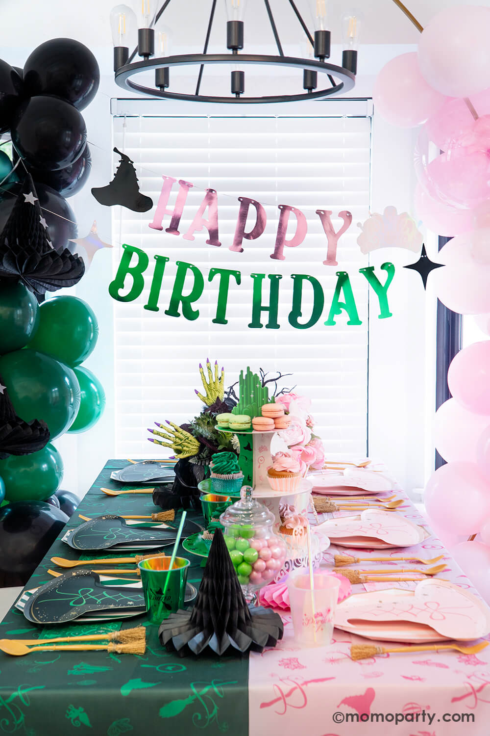 A spellbinding Wicked birthday party set up by Momopartys featuring various Wicked themed party supplies and decorations that represent two worlds or Glinda and Elphaba including pink and black witch hat shaped plates, two-toned Emerald City cupcake stand, Wicked party cups, napkins, witch broom utensil set and Wicked witch hat honeycombs on the green and pink two-tone tablecloth. With Wicked floral arrangement and treats on the table, balloon garlands and happy birthday banner hung above as the backdrop.