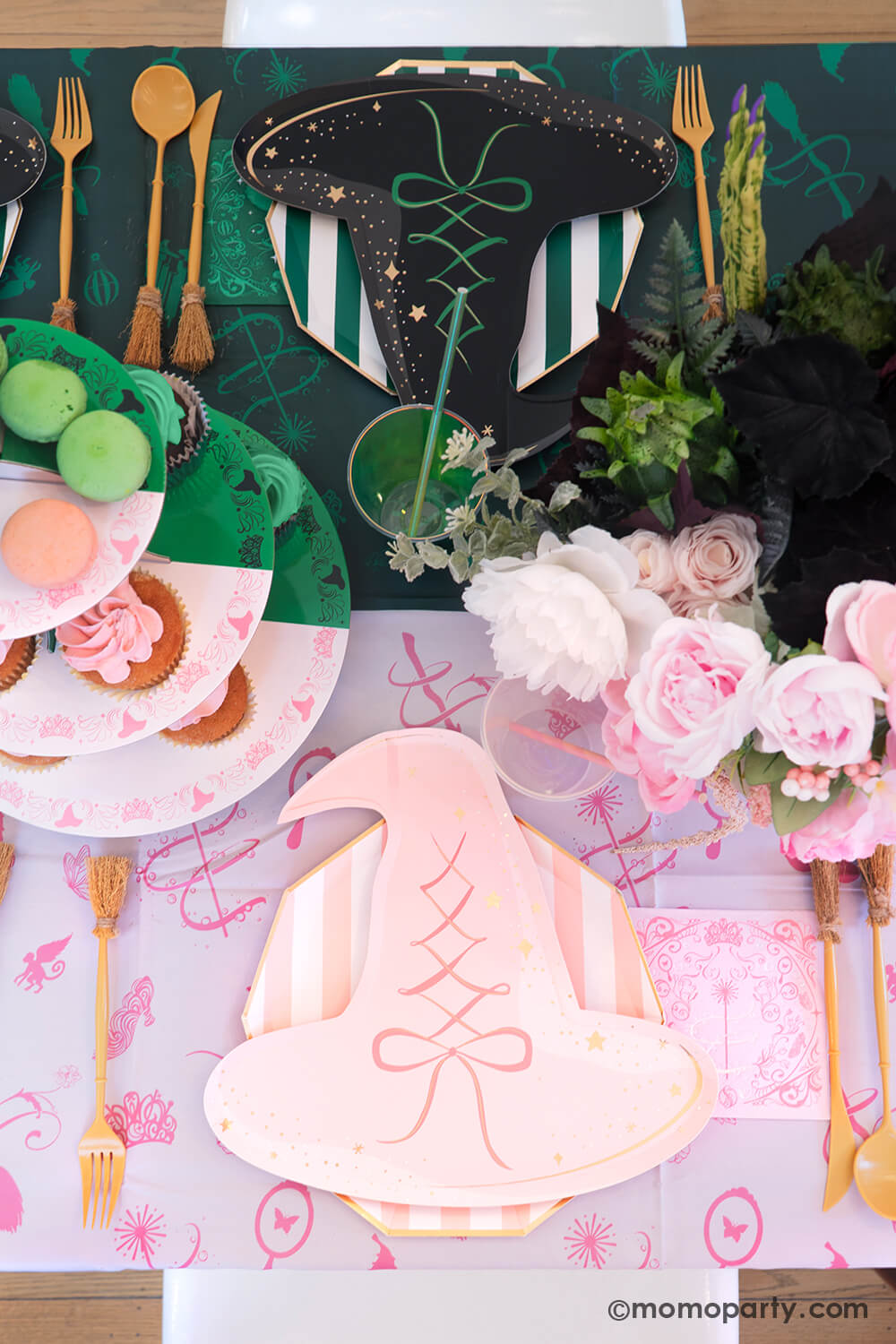 A Wicked themed party table setting with various of Momopartys's Wicked themed party supplies including witch hat shaped plates in pink and black, napkins inspired by Glinda and Elphaba, witch broom shaped utensils, and Wicked Emerald City 3 tier cupcake stand on the pink and green two-tone Wicked tablecloth. With Wicked inspired floral bouquet, it makes a spellbinding Halloween celebration in this spooky season!