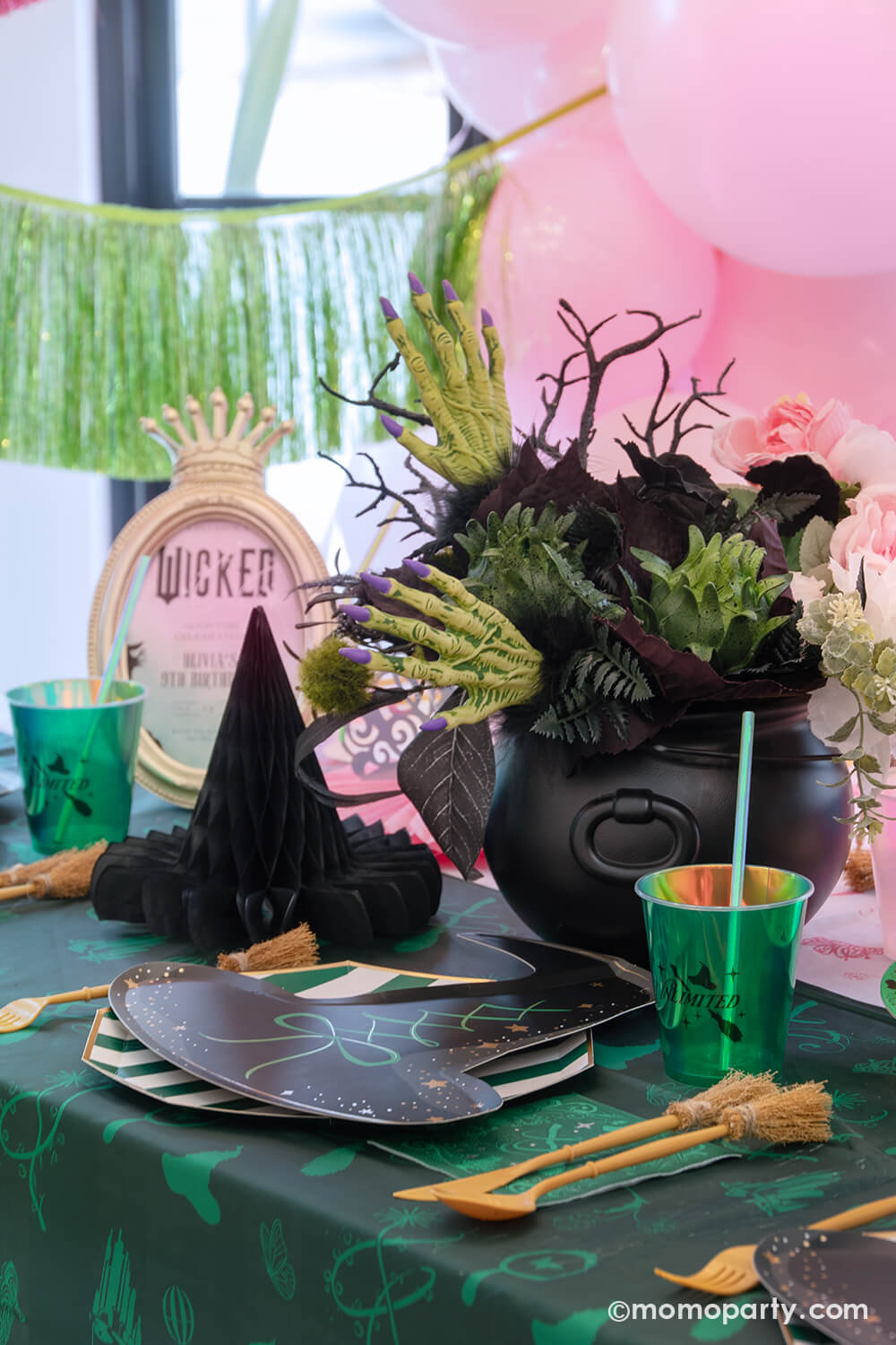 A Wicked themed party table setting with various of Momopartys's Wicked themed party supplies including the wicked witch hat shaped plates, emerald green striped dinner plates, the Wicked green napkins inspired by Elphaba, witch broom shaped utensil set, and wicked green party cups with green iridescent straws on the green and pink two-tone Wicked tablecloth. With Wicked inspired floral bouquet and witch hat honeycombs, it's perfect for a spellbinding celebration.