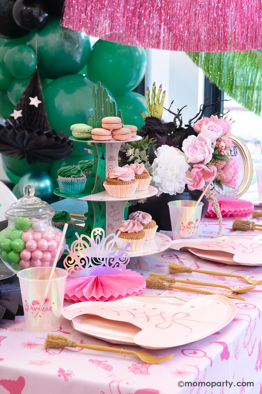 A Glinda inspired Wicked themed party table setting featuring various Wicked themed party supplies and decorations from Momopartys including Glinda pink tiaras, pink witch hat shaped plates, pink and green two-toned Wicked Emerald City cupcake stand, Glinda iridescent party cups, witch broom utensil set and Wicked witch hat honeycombs on the pink tablecloth. With green and pink floral bouquet and treats, this makes a mystical yet whimsical table for a Wicked themed party this spellbinding season!