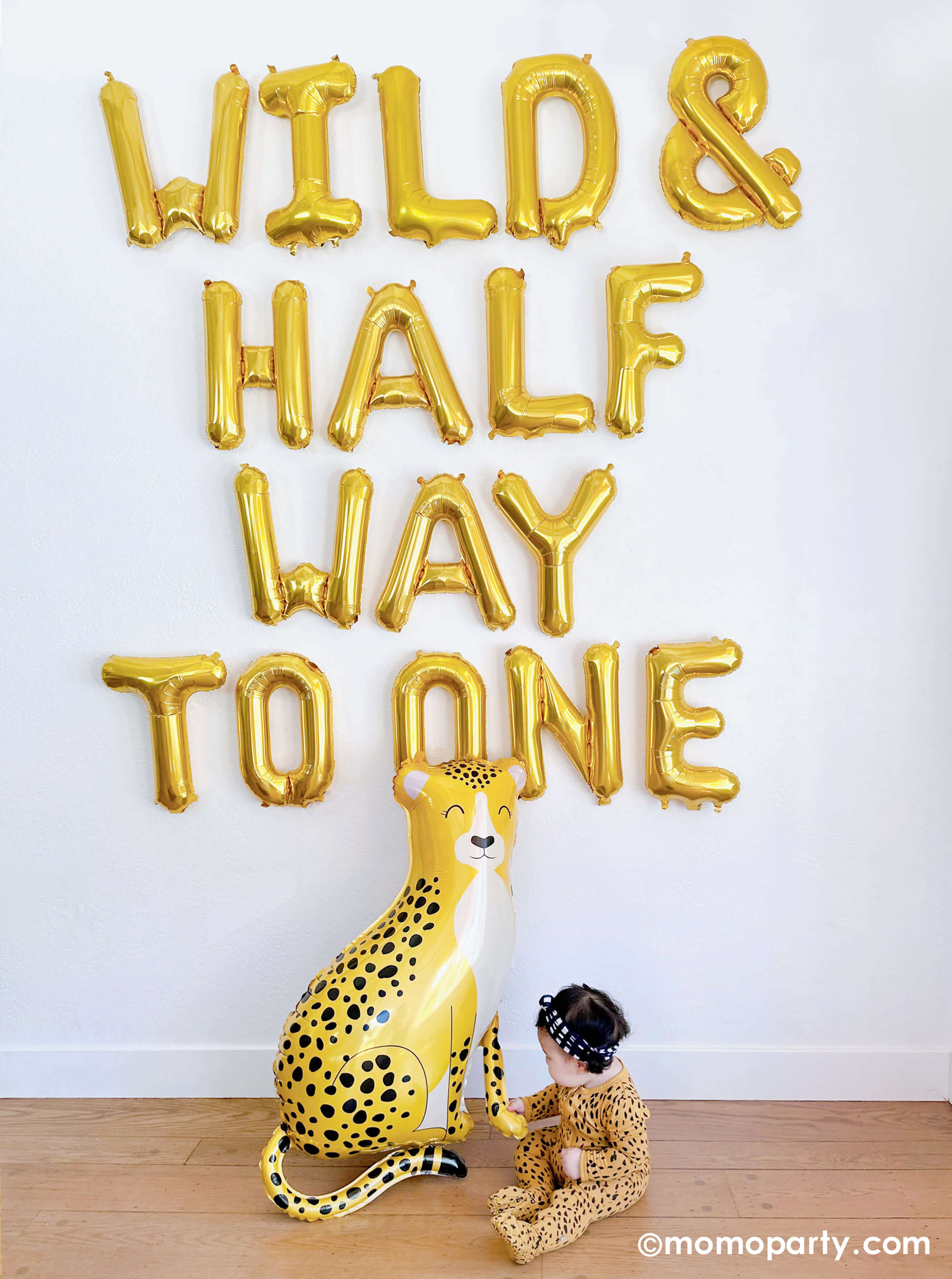 A balloon wall featuring Momopartys's "Wild & Half Way To One" 16" Mylar Letter Balloons by Northstar Letter Balloons. At the bottom of the wall sits Momopartys's baby cheetah shaped foil balloon by Betallic Balloons and a 6-month baby girl in her cheetah print onese reaching out her hand to hold the cheetah, all makes a great inspiration for an adorable baby's half birthday party.
