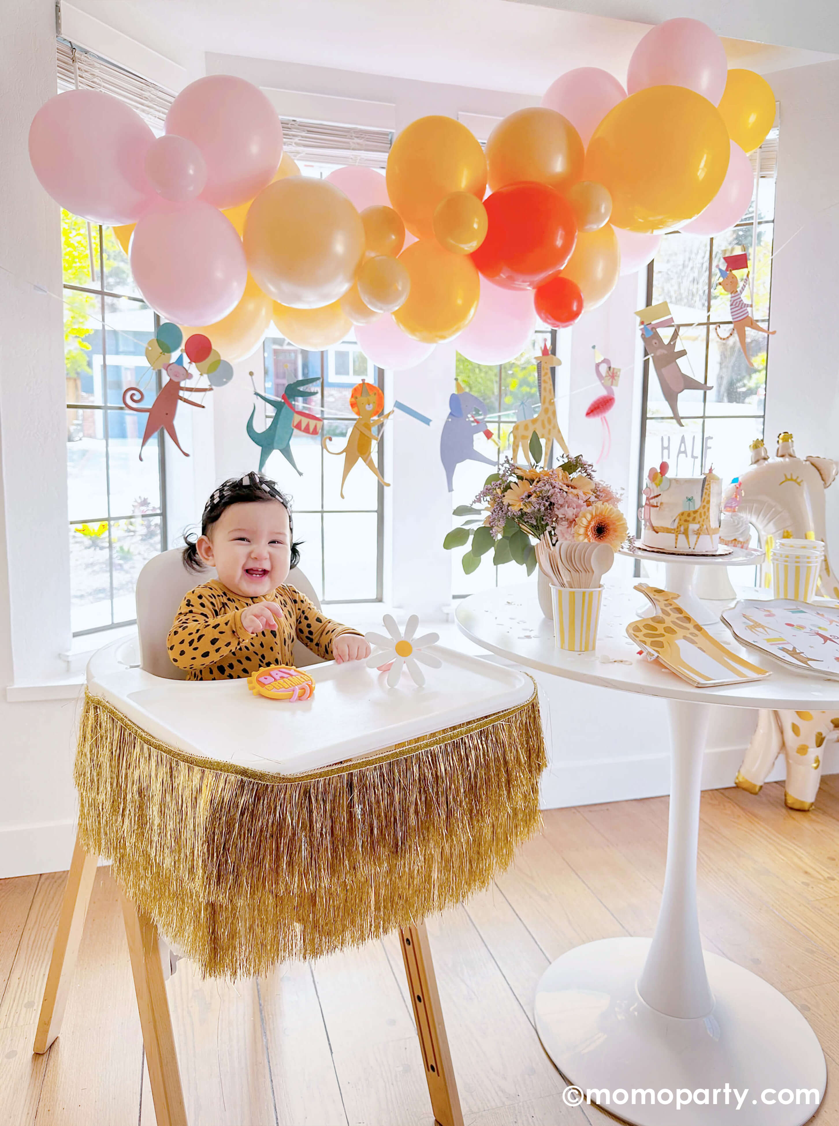 A happy baby girl's 6 months animal themed birthday party featuring Momopartys's animal parade birthday garland featuring a lion, an elephant, an alligator, a giraffe, along with a festive balloon garland in a warm color tone hanging above. On the dessert table, there are Meri Meri's animal parade dinner plates, giraffe shaped plates and cupcakes dressed with the safari animal toppers, next to the table there's a giraffe shaped foil balloon. One the left a highchair is decorated with a gold tinsel garland.