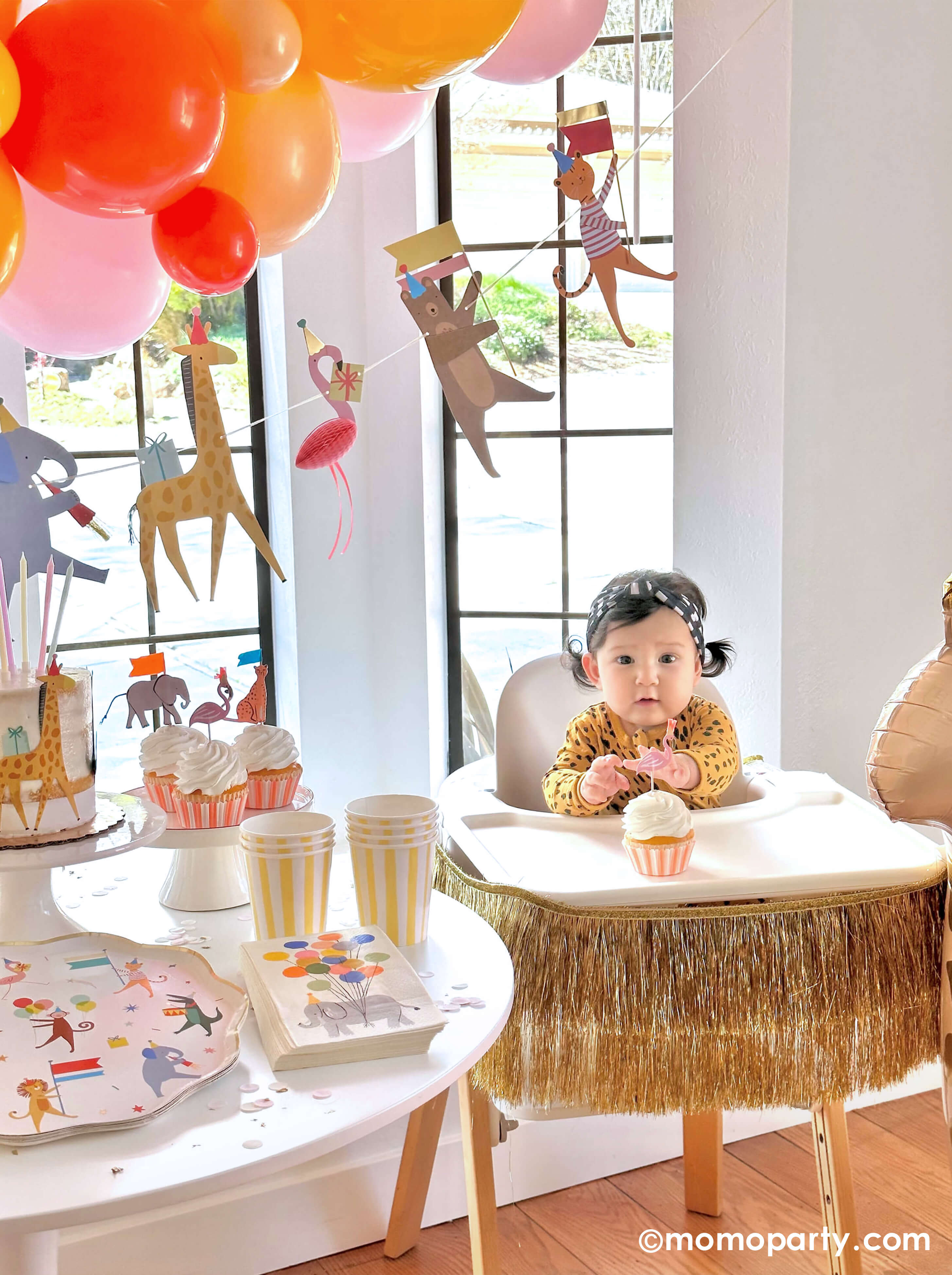 A cute baby girl wearing animal pattern onesie, sitting on a highchair in her 6 months animal themed half birthday party, reaching a cupcake decorated with Meri Meri Safari Animals Cupcake Kit, next to a party table featuring Momopartys's Animal Parade Dinner plate, Rifle Paper Co's party animal guest napkins, a cake decorated with  , along with light yellow striped cups and yellow wooden utensil.  it makes a great party inspiration for kid's animal, safari, carnival themed birthday party.