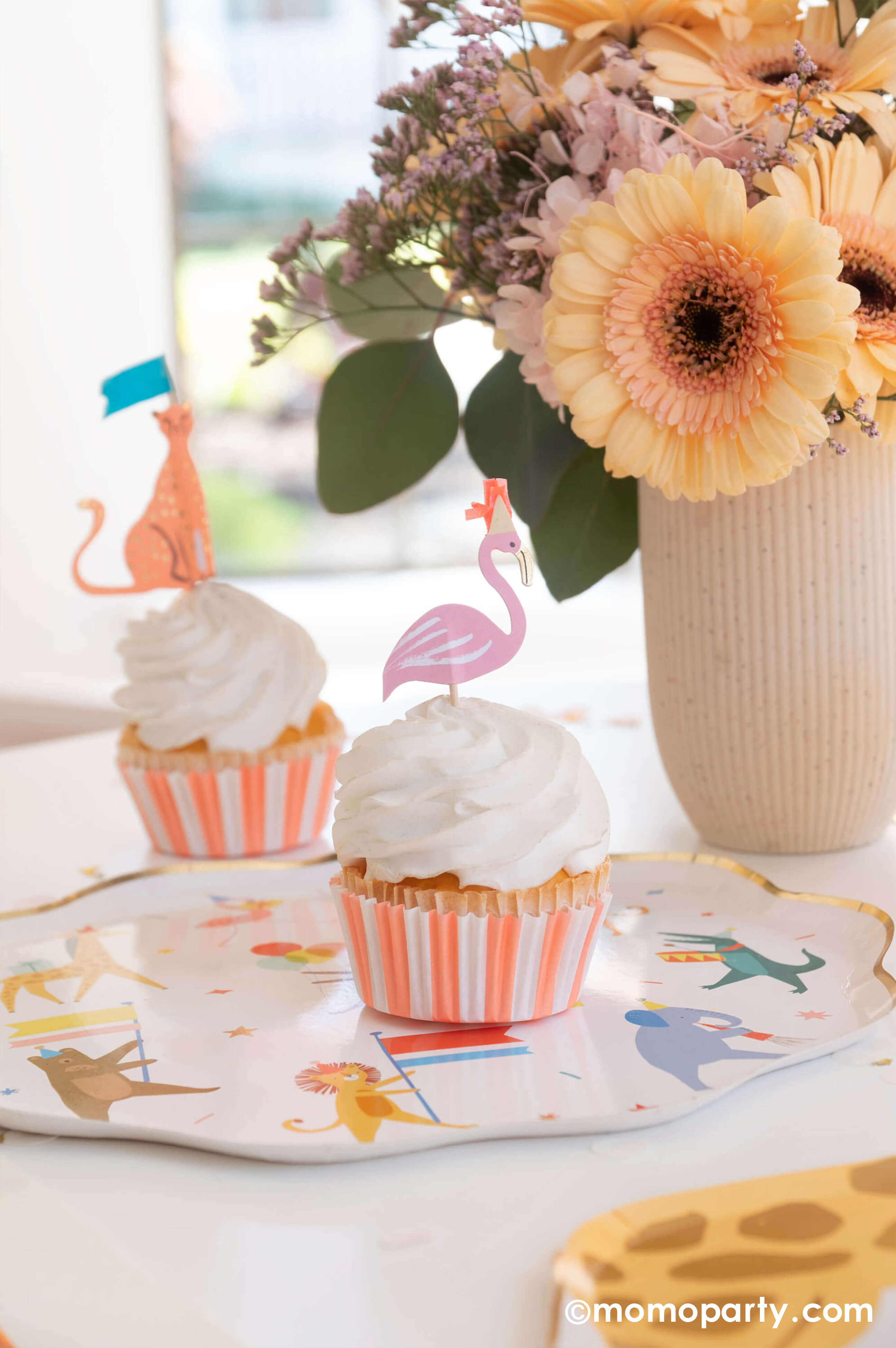 A kid's animal themed party table featuring Momopartys's Animal Parade Dinner plate, a giraffe shaped plate for cakes, and adorable safari animal cupcake toppersby Meri Meri, and Rifle Paper Co's party animal guest napkins with a cute illustration of an elephant with a party hat and bunch of festive balloons, along with light yellow striped cups and wooden utensil, it makes a great party inspiration for kid's animal, safari, carnival themed birthday party.