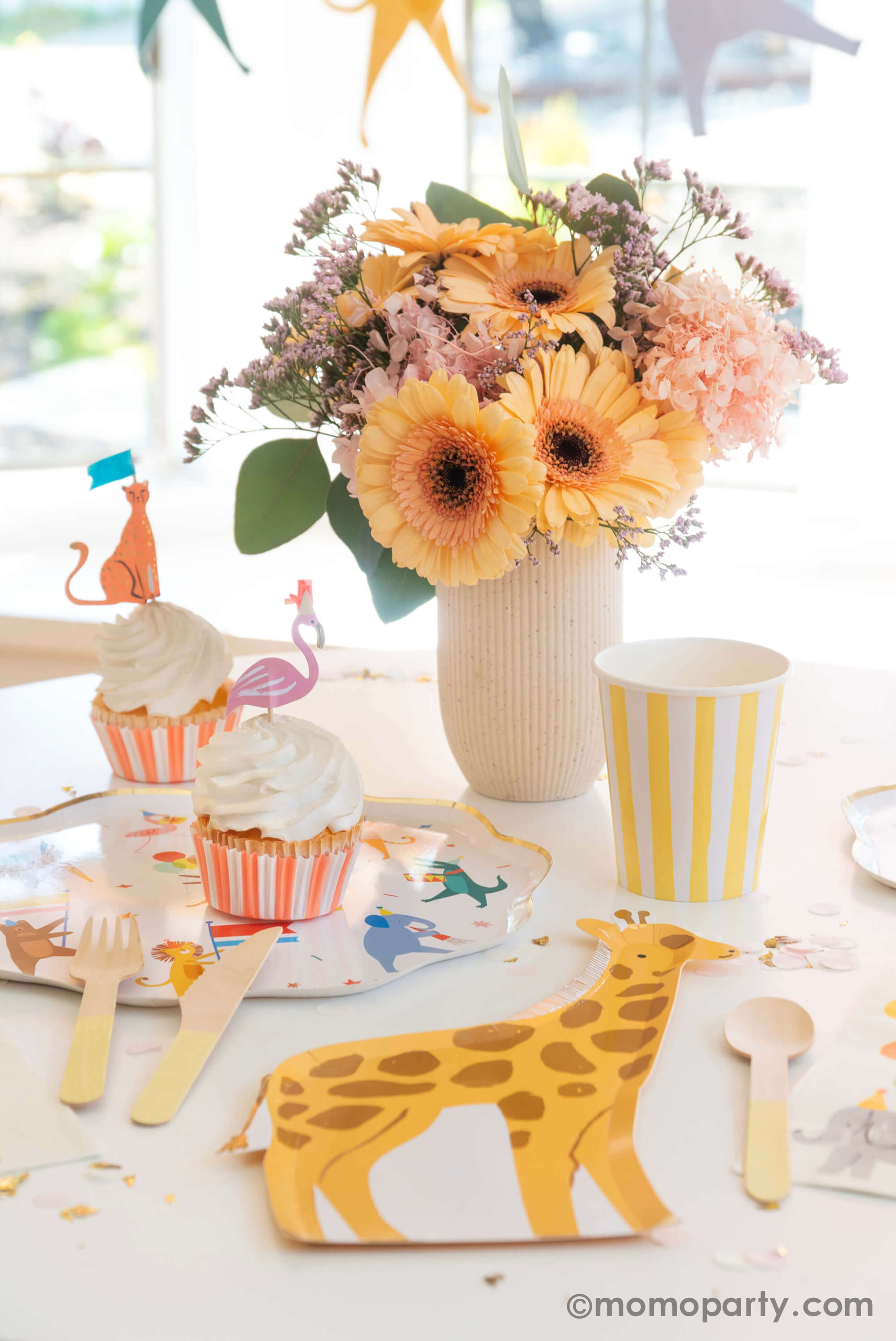 A kid's animal themed party table featuring Momopartys's Animal Parade Dinner plate, a giraffe shaped plate for cakes, and adorable safari animal cupcake toppersby Meri Meri, and Rifle Paper Co's party animal guest napkins with a cute illustration of an elephant with a party hat and bunch of festive balloons, along with light yellow striped cups and wooden utensil, it makes a great party inspiration for kid's animal, safari, carnival themed birthday party.