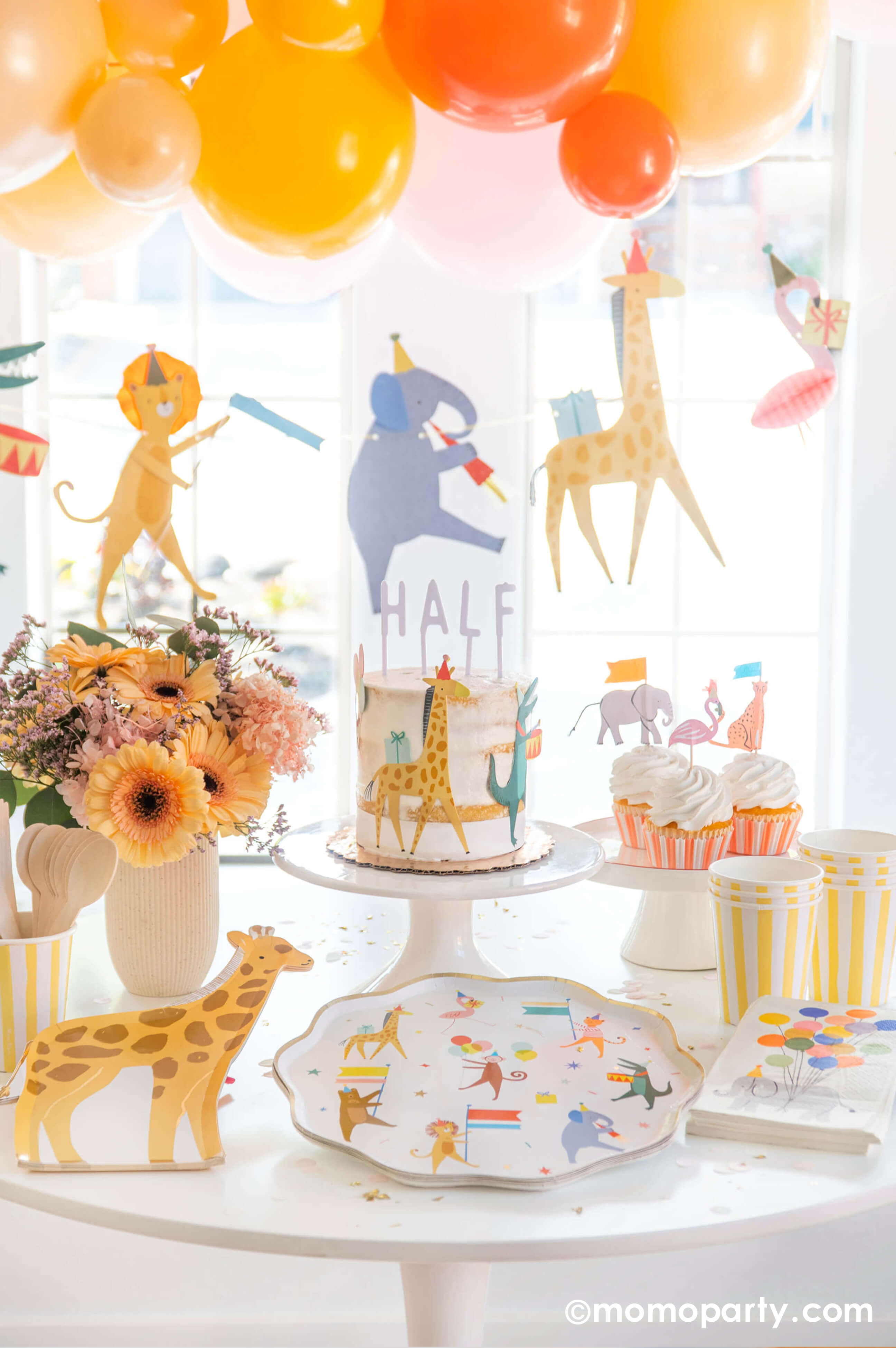 A kid's animal themed party table featuring Momopartys's Animal Parade Dinner plate, giraffe shaped plates, light yellow striped cups and safari animal cake toppers by Meri Meri, along with a beautiful spring flower arrangement as the centerpiece and a fun animal parade birthday garland featuring a lion, an elephant, a giraffe, a flamingo, and a festive balloon garland in a warm color tone in the back, it makes a great party inspiration for kid's animal, safari, carnival themed birthday party.