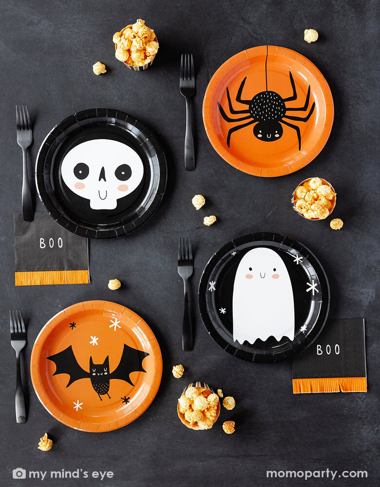 A kid's friendly Halloween party tablescape features Momopartys's classic Halloween party supplies including the friendly black and orange round plates with adorable character illustrations like a bat, a skull, a ghost and bat on them , the boo fringe small napkins, and the orange and black striped food cups filled with popcorn. This simple and modern place setting provide an effortless yet festive idea for celebrating Halloween with kids this spooky season.