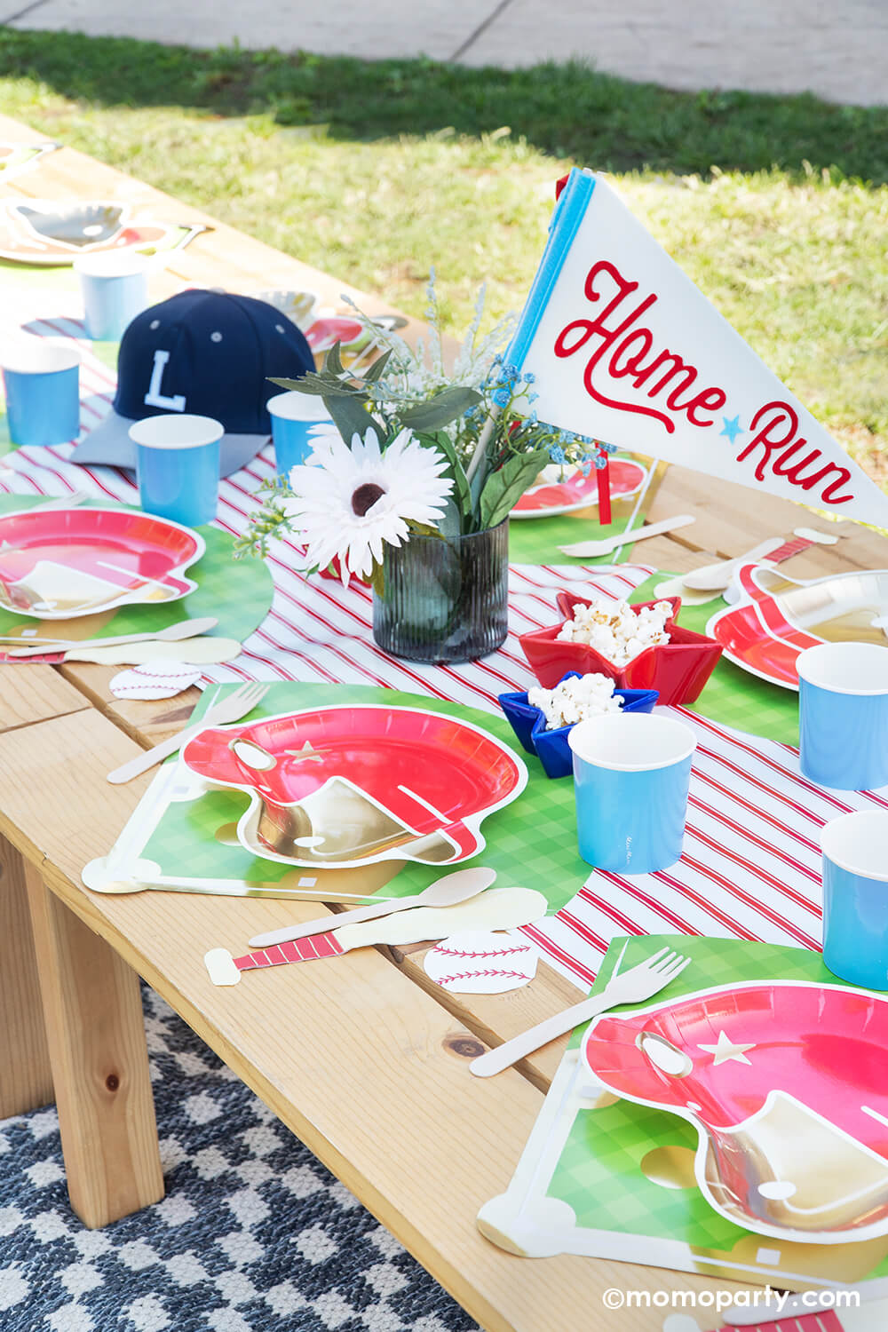A kid's baseball themed birthday party picnic table setting styled by Momopartys featuring various baseball themed tablewares from Momopartys including baseball helmet shaped plate, cornflower tumbler cups, baseball shaped napkins on the baseball diamond placemats with the red pinstriped runner underneath. In the center there's a beautiful flower bouquet with the "Home Run" felt party pennant and some star shaped trays in blue and red filled with popcorn.
