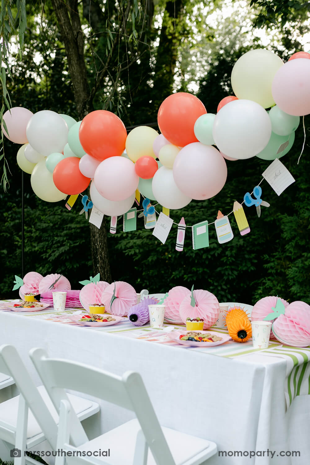 A backyard pastel themed back to school party table set up and decoration featuring Momopartys's back to school themed party goods including back to school party garland, pink apple honeycombs and rainbow pastel pencil honeycombs as the centerpiece, ABC alphabet plates, party cups and napkins. With a pastel themed balloon garland hung above the table, it makes an adorable celebration for kid's first day of school.