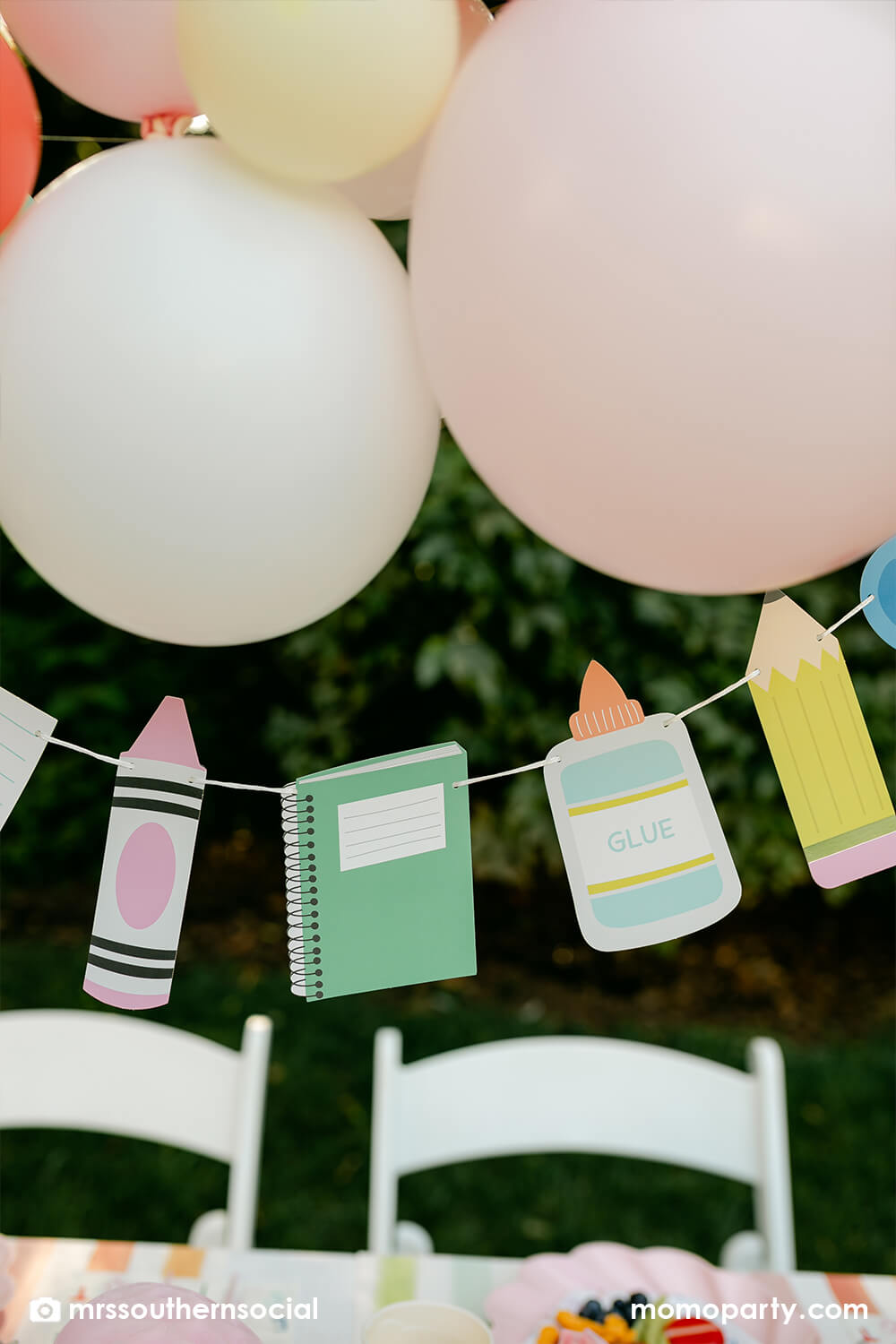 A backyard pastel themed back to school party decorated with Momopartys's back to school themed party goods including back to school party garland featuring school supply pennants including pencils, glue bottles, crayons, notebooks,  and more. With a pastel themed balloon garland hung above the table, it makes an adorable celebration for kid's first day of school.