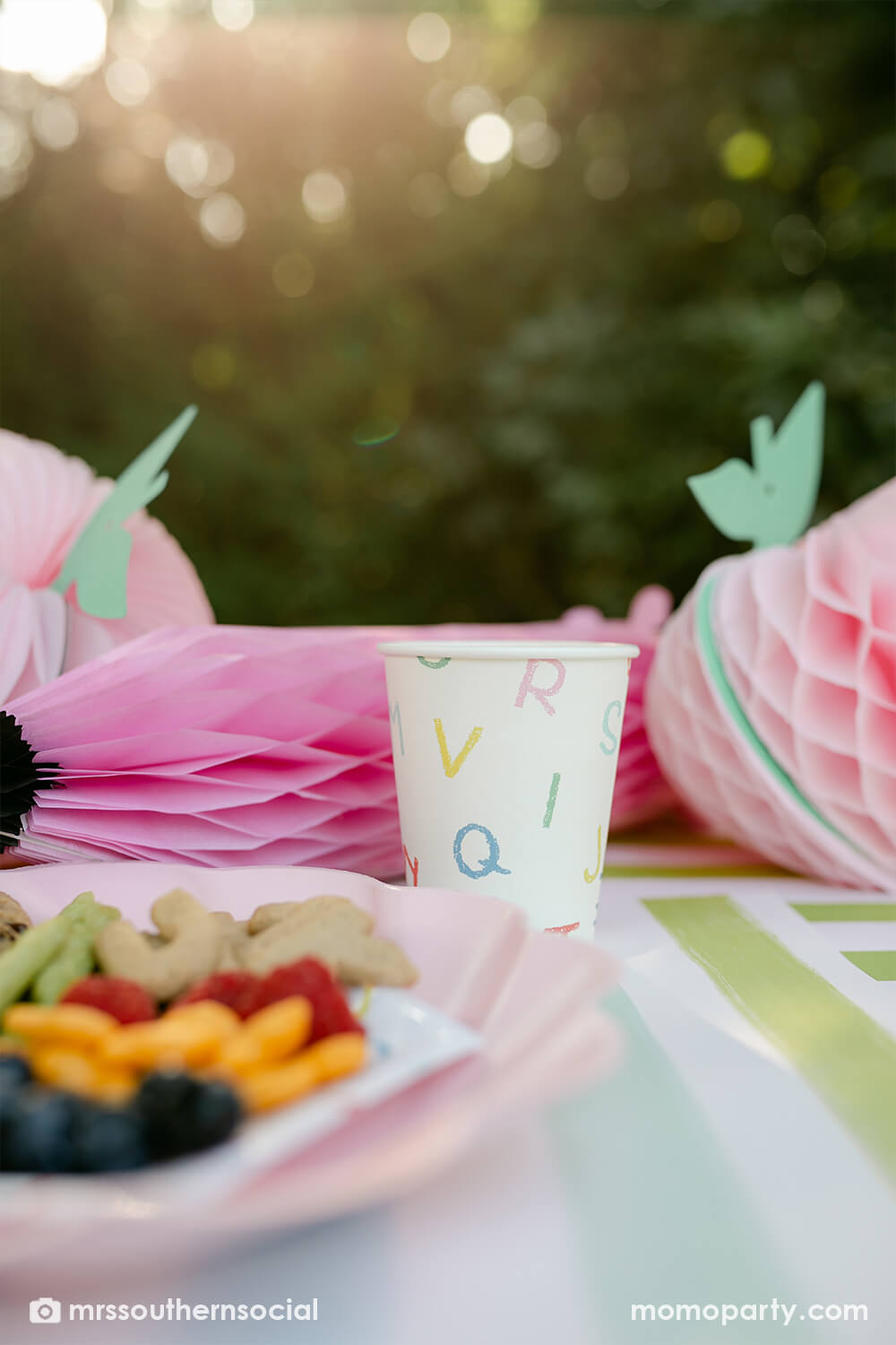 A backyard pastel back to school party table featuring Momopartys's back to school party supplies including ABC alphabet plates, napkins, party cups by Merrilulu. With pastel rainbow striped placemats and pastel rainbow pencil honeycombs and pink apple honeycombs, all together makes an adorable tablescape for kid's first day of school celebration.