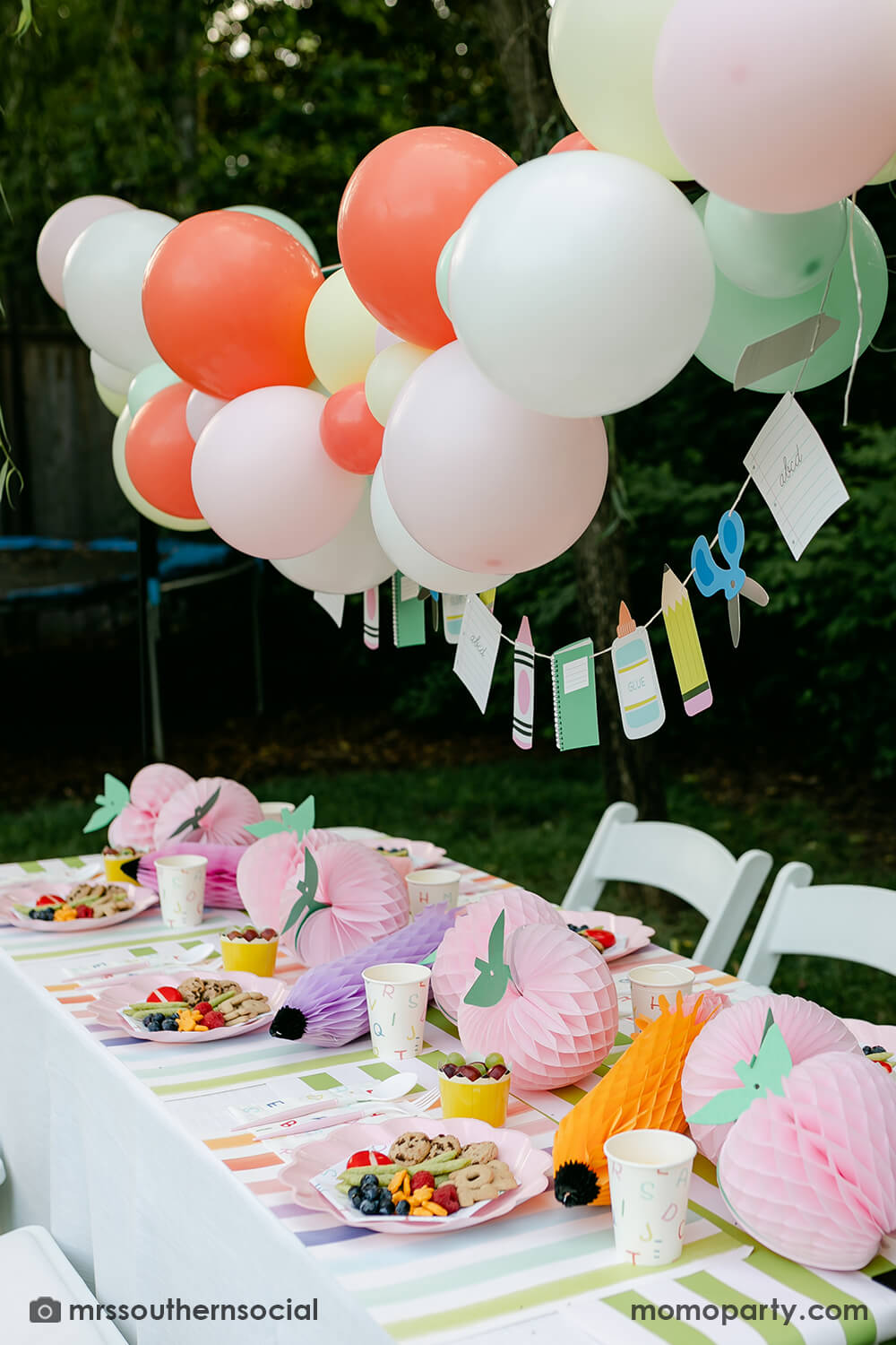 A backyard pastel themed back to school party table set up and decoration featuring Momopartys's back to school themed party goods including back to school party garland, pink apple honeycombs and rainbow pastel pencil honeycombs as the centerpiece, ABC alphabet plates, party cups and napkins. With a pastel themed balloon garland hung above the table, it makes an adorable celebration for kid's first day of school.