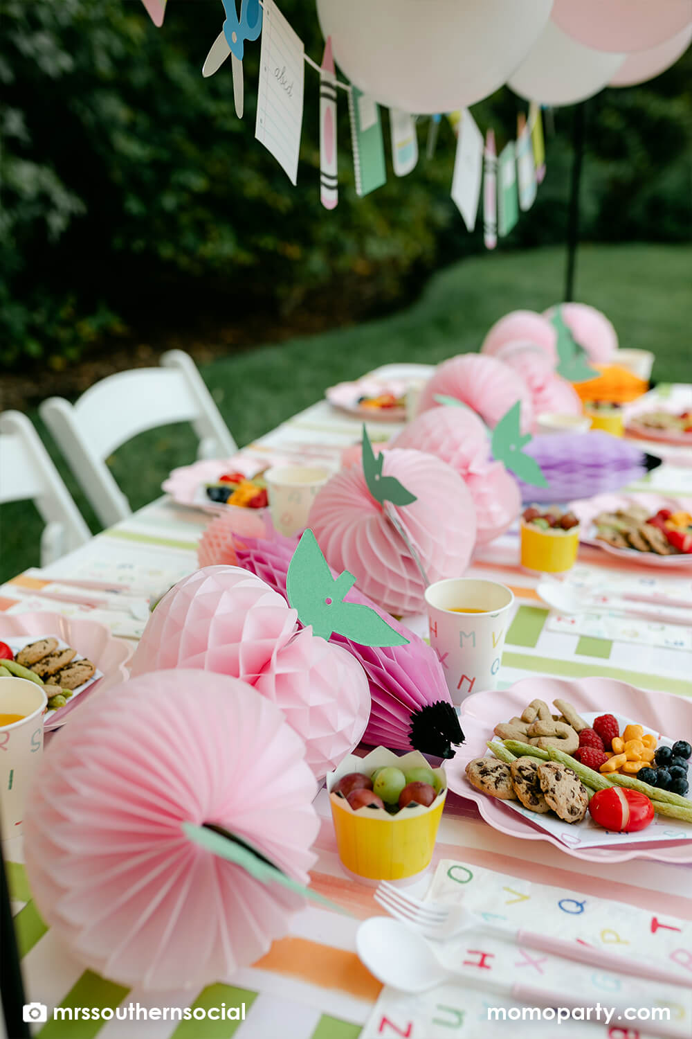 A backyard pastel themed back to school party table set up and decoration featuring Momopartys's back to school themed party goods including back to school party garland, pink apple honeycombs and rainbow pastel pencil honeycombs as the centerpiece, ABC alphabet plates, party cups and napkins. With kids snacks including ABC alphabet crackers, fruits, cheese, it gives great ideas and inspo for celebrating kid's first day of school.