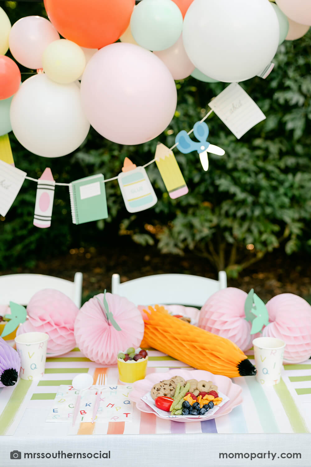A backyard pastel themed back to school party table set up and decoration featuring Momopartys's back to school themed party goods including back to school party garland, pink apple honeycombs and rainbow pastel pencil honeycombs as the centerpiece, ABC alphabet plates, party cups and napkins. With a pastel themed balloon garland hung above the table, it makes an adorable celebration for kid's first day of school.