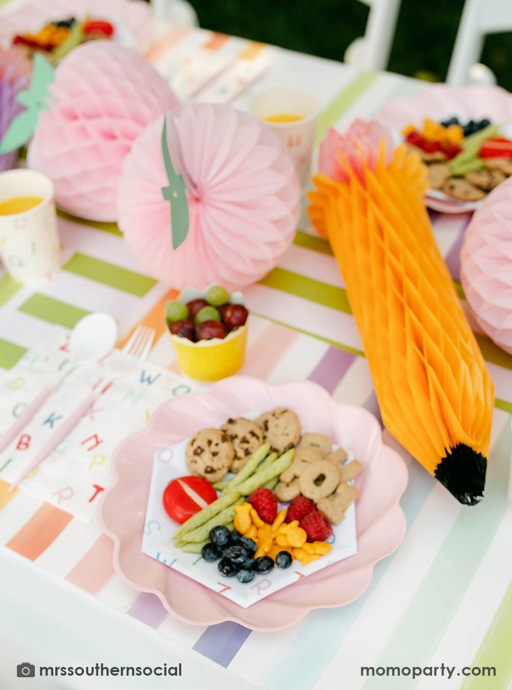 A pastel back to school party table featuring Momopartys's back to school party supplies including ABC alphabet plates, napkins party cups, and pencil shaped food cups. One the plates are kids snack ideas including fruits, cheeses, goldfish, ABC alphabet crackers. With pastel rainbow striped placemats and pastel rainbow pencil honeycombs and pink apple honeycombs, all together makes an adorable tablescape for kid's first day of school celebration.