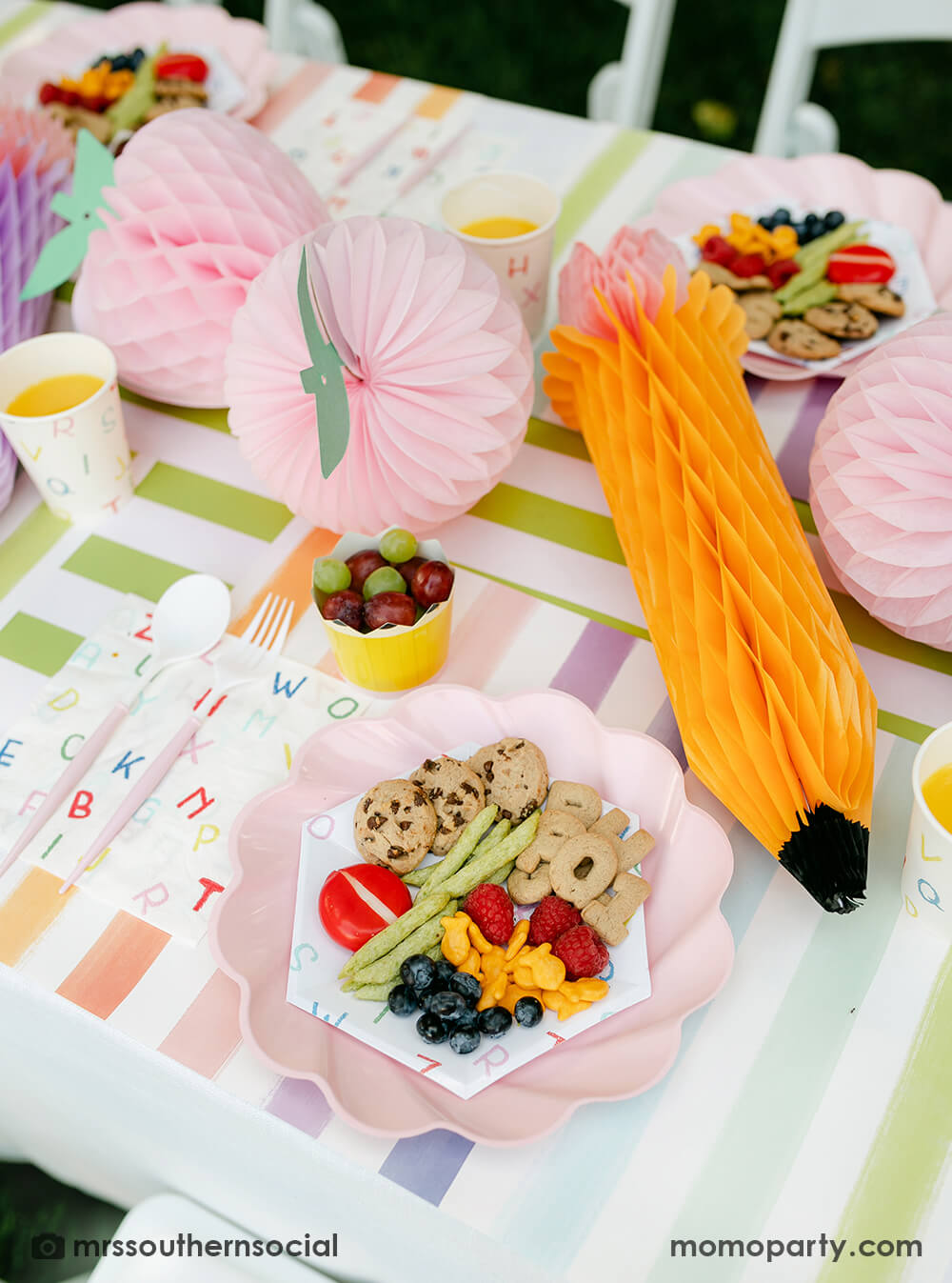 A pastel back to school party table featuring Momopartys's back to school party supplies including ABC alphabet plates, napkins party cups, and pencil shaped food cups. On the plates are kids snack ideas including fruits, cheeses, goldfish, ABC alphabet crackers. With pastel rainbow striped placemats and pastel rainbow pencil honeycombs and pink apple honeycombs, all together makes an adorable tablescape for kid's first day of school celebration.