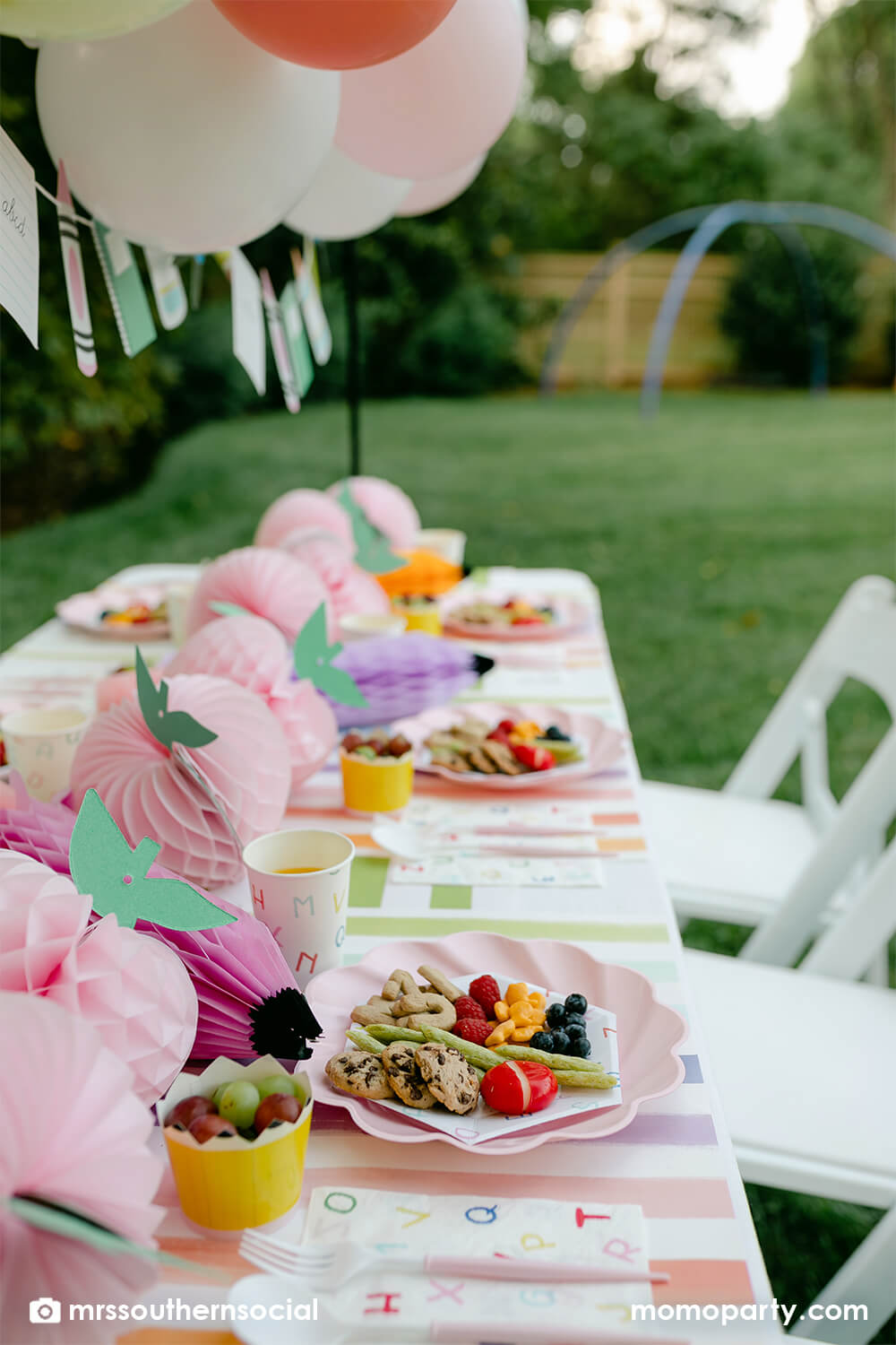 A backyard pastel themed back to school party table set up and decoration featuring Momopartys's back to school themed party goods including back to school party garland, pink apple honeycombs and rainbow pastel pencil honeycombs as the centerpiece, ABC alphabet plates, party cups and napkins. With kids snacks including ABC alphabet crackers, fruits, cheese, it gives great ideas and inspo for celebrating kid's first day of school.
