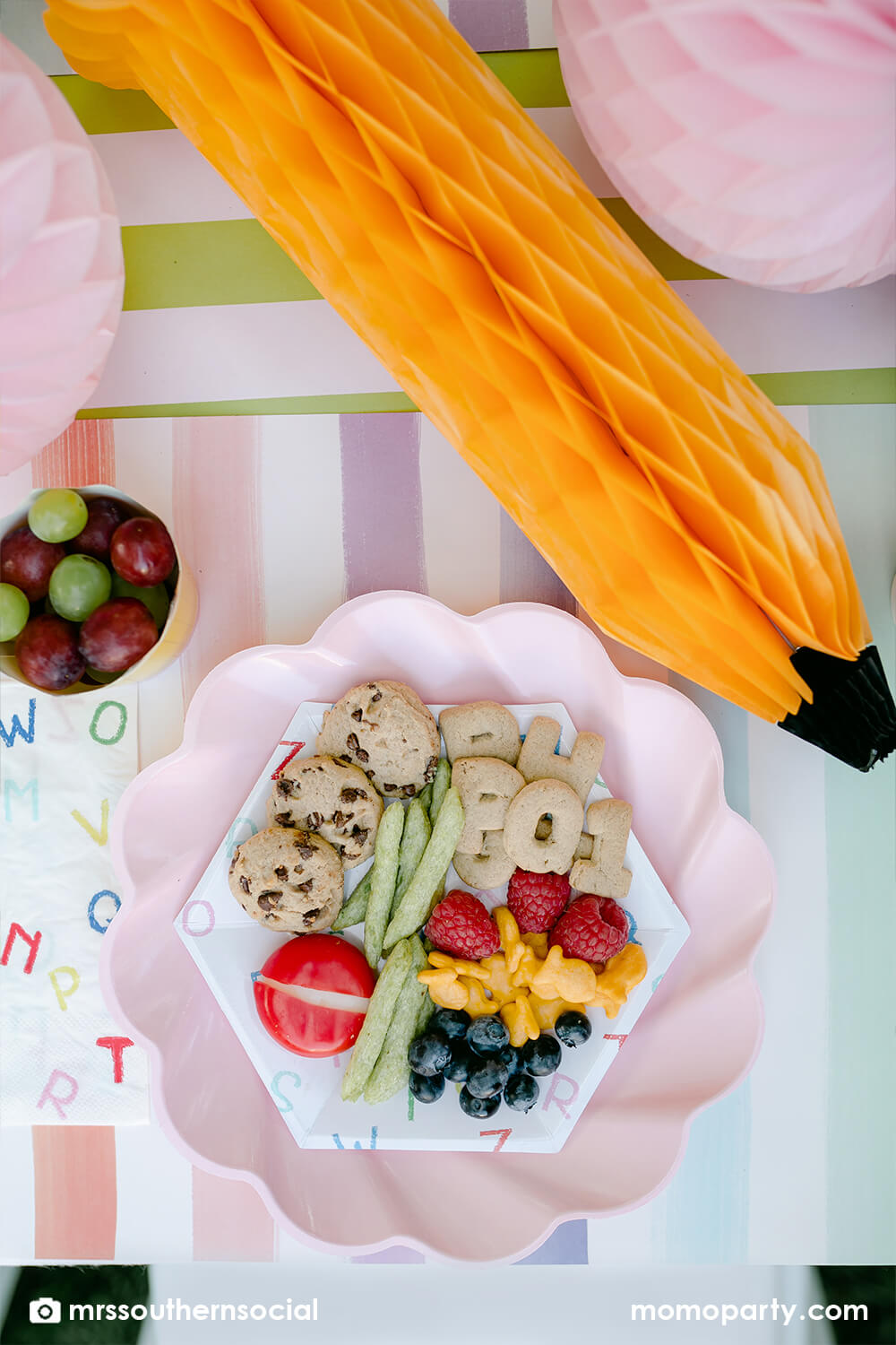 A pastel back to school party table featuring Momopartys's back to school party supplies including ABC alphabet plates, napkins party cups, and pencil shaped food cups. One the plates are kids snack ideas including fruits, cheeses, goldfish, ABC alphabet crackers. With pastel rainbow striped placemats and pastel rainbow pencil honeycombs and pink apple honeycombs, all together makes an adorable tablescape for kid's first day of school celebration.