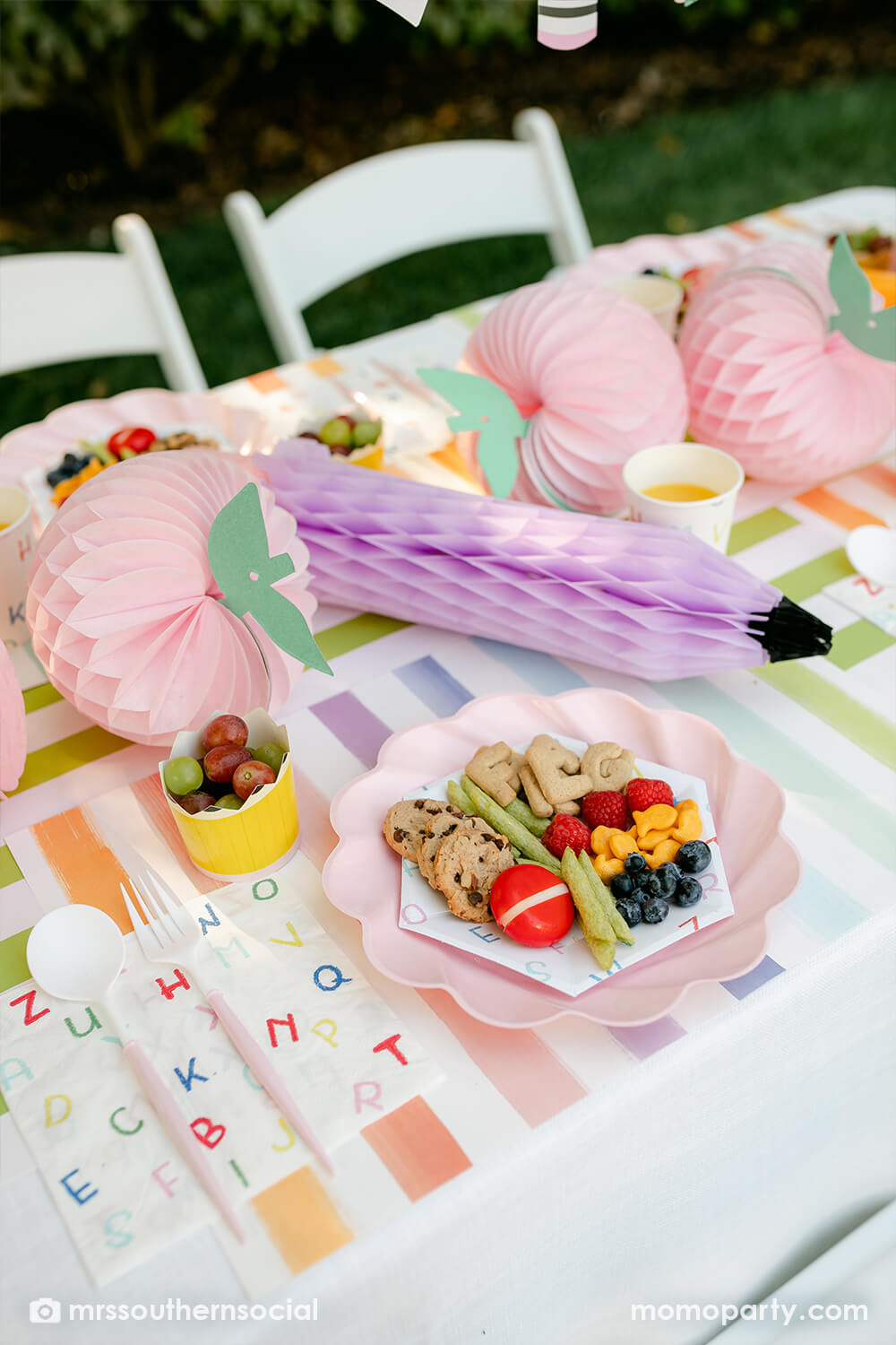 A pastel back to school party table featuring Momopartys's back to school party supplies including ABC alphabet plates, napkins party cups, and pencil shaped food cups. On the plates are kids snack ideas including fruits, cheeses, goldfish, ABC alphabet crackers. With pastel rainbow striped placemats and pastel rainbow pencil honeycombs and pink apple honeycombs, all together makes an adorable tablescape for kid's first day of school celebration.