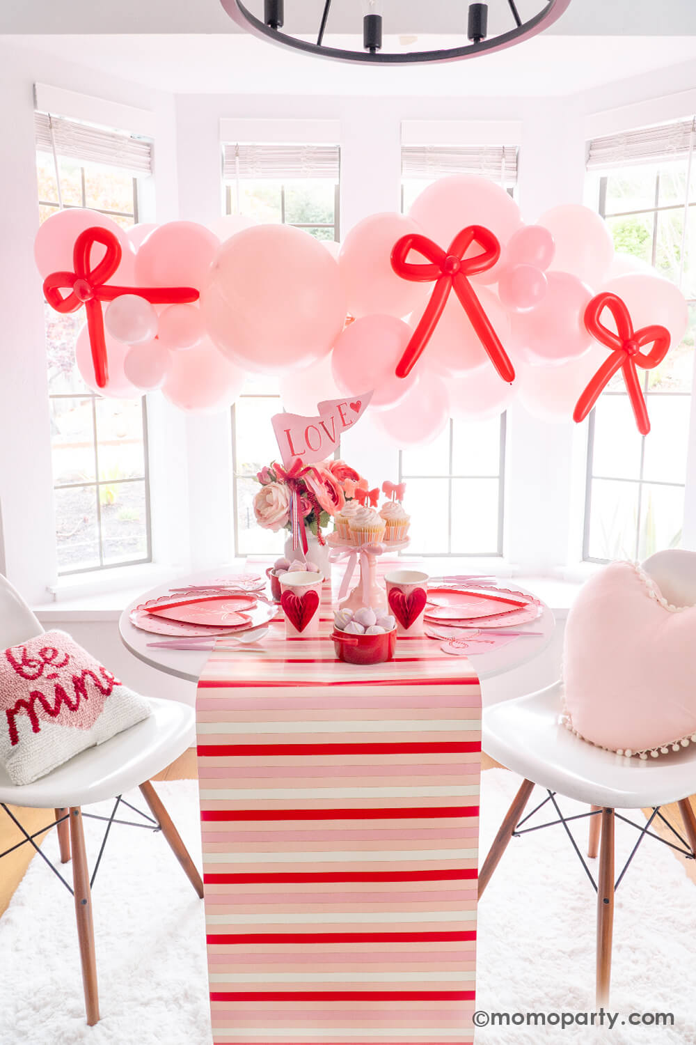 Tying the Knot of Love: A Bow-Themed Valentine's Day Party Set up by Momopartys. A pink balloon garland with a red bow-shaped balloon hangs above the Bow-tastic Table, adorned with Meri Meri Heart Pattern Dinner Plates, Bow Plates, matching napkins, Honeycomb Heart Cups, Blush and White Cutlery Set, Pink Bow Candles on cupcakes, and a Love Party Pennant gracing the flower vase. All elegantly arranged on a red-striped paper runner. A cute idea for your Valentine's celebration or Galentine's Day with besties