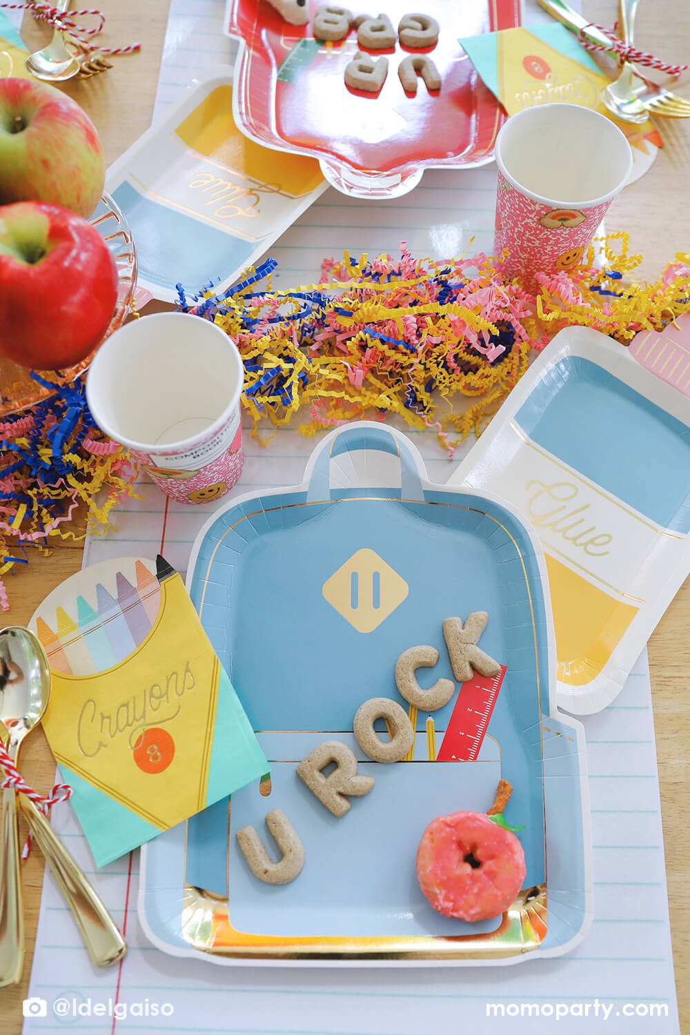 A festive back to school party tablescape featuring Momopartys's school themed party supplies including backpack shaped plate set in three different colors, glue bottle shaped plates, crayon box shaped napkins, pink notebook inspired party cups and giant notebook placemats. On the plates, there are alphabet shaped cookies spelling "U Rock" and apple shaped mini donuts, with apples stacked as the centerpiece and some fun confetti, this makes a perfect first day of school celebration for kids.