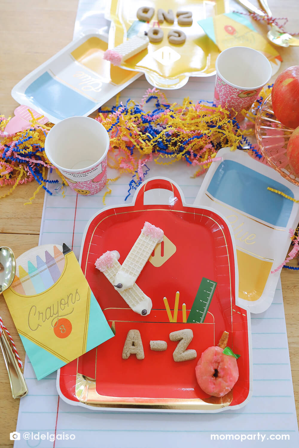A festive back to school party tablescape featuring Momopartys's school themed party supplies including backpack shaped plate set in three different colors, glue bottle shaped plates, crayon box shaped napkins, pink notebook inspired party cups and giant notebook placemats. On the plates, there are alphabet shaped cookies and pencil shaped wafer cookies, with apples stacked as the centerpiece and some fun confetti, this makes a perfect first day of school celebration for kids.