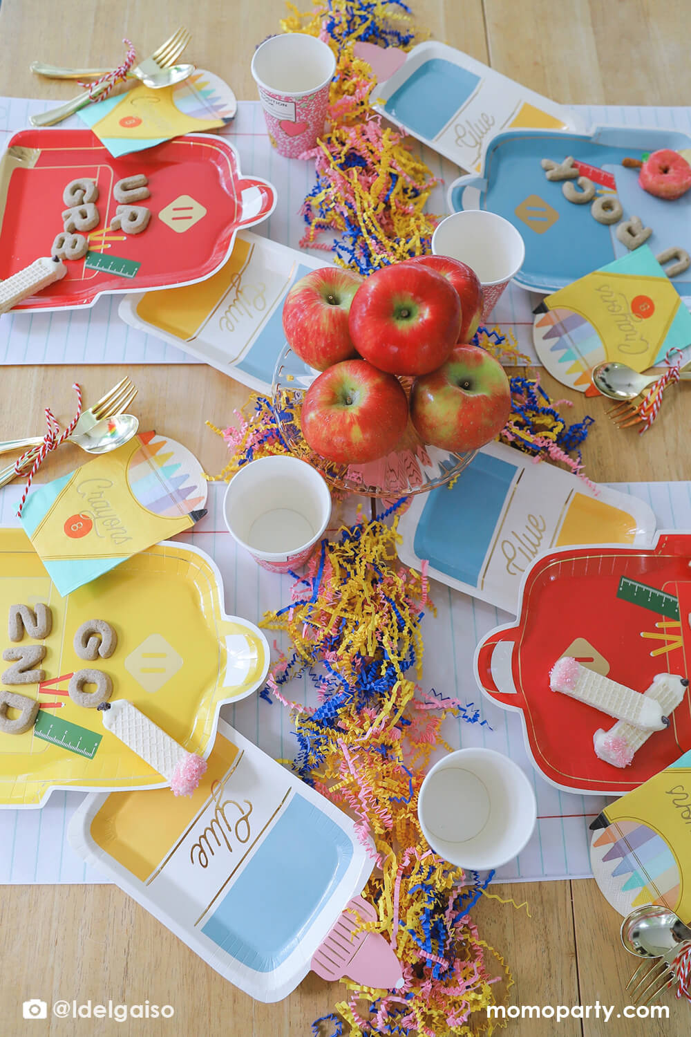 A overhead shot of a festive back to school party table featuring Momopartys's school themed party supplies including backpack shaped plate set in three different colors of red, blue and yellow, glue bottle shaped plates, crayon box shaped napkins, pink notebook party cups and giant notebook placemats. On the plates, there are alphabet shaped cookies and pencil shaped wafer cookies, with apples stacked as the centerpiece and some fun confetti, this makes a perfect first day of school celebration for kids.