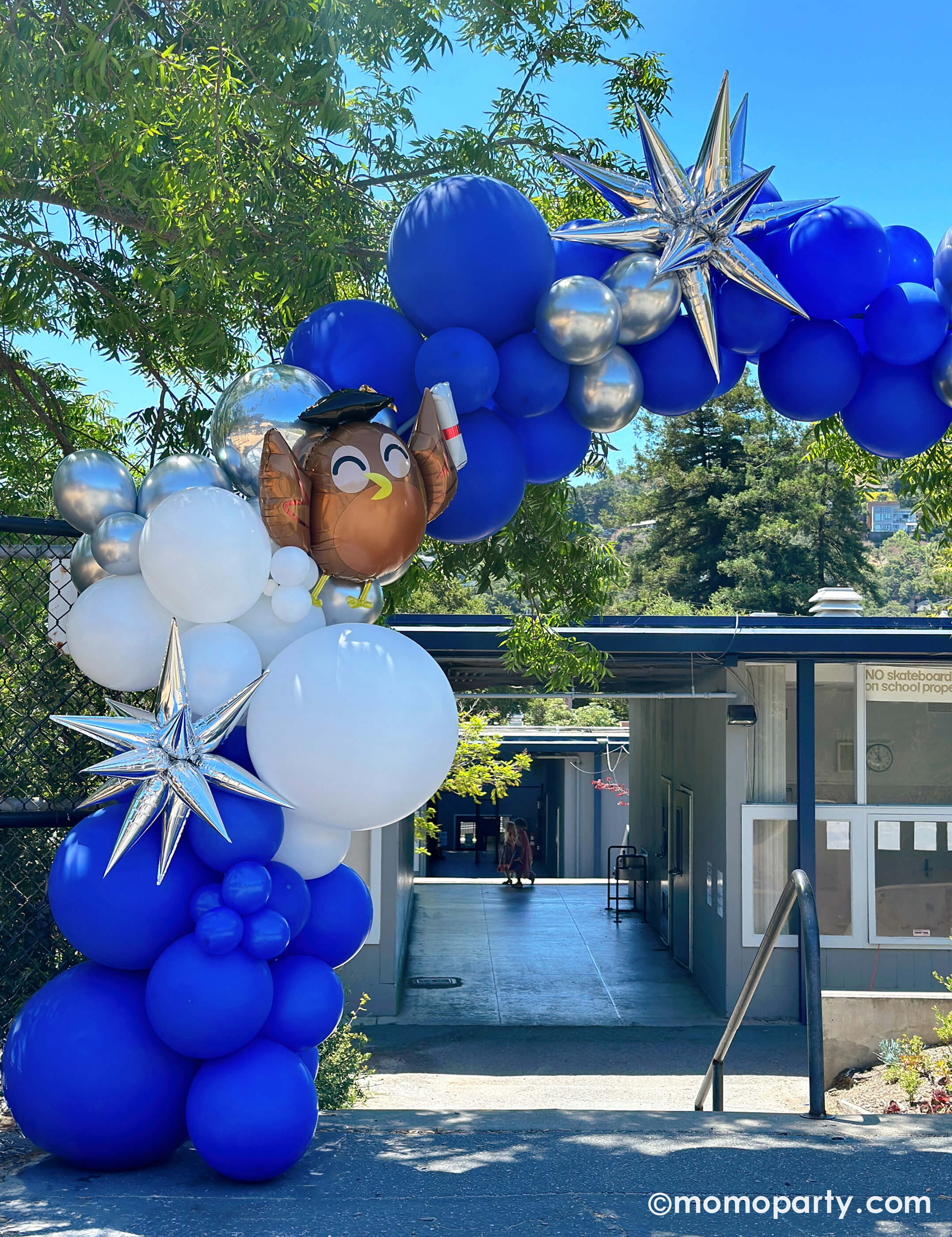 Elementary School Graduation Party by Momopartys. Featuring a stunning balloon arch in school colors of blue, white, and silver, accented with Graduate Owl Shaped Foil Balloons and a 12 Point Silver Starburst Foil Balloon in front of the school staircase. This arch is a big hit, serving as a graduation gate for students to pass through and a photo backdrop to capture last day of school memories.