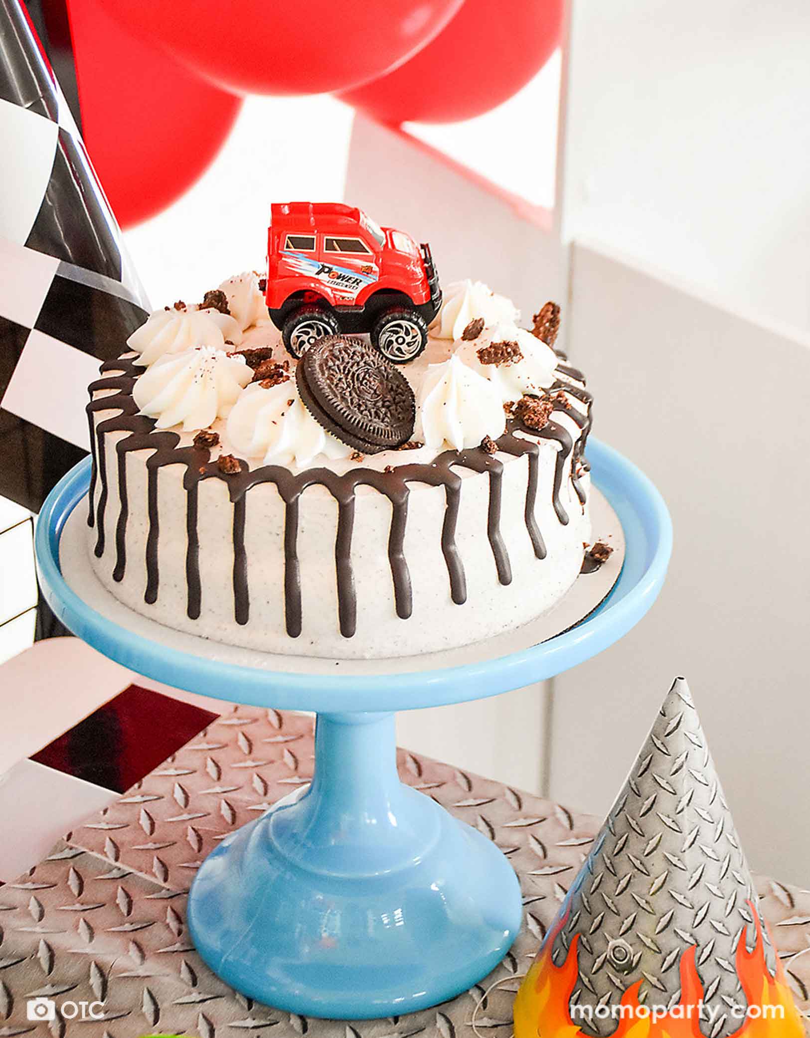 A monster truck themed birthday party table with a Oreo ice cream birthday cake decorated with Momopartys's red pull-back toy monster truck toy. On the table there's a party hat and a tablecloth in the steel texture design, along with race car checkered flags and red balloons, a fun and exciting monster truck birthday party awaits!