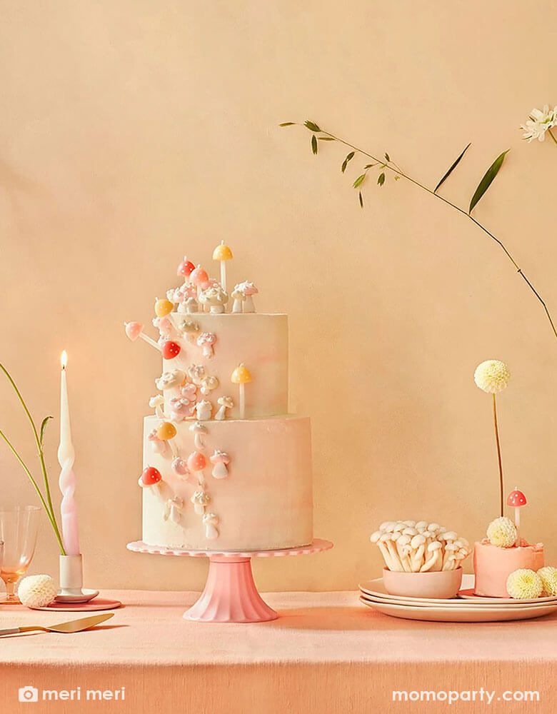 A light pink buttercream cake decorated with Momopartys's 3D mushroom birthday candles by Meri Meri on a pink cake stand. On the table there are also other desserts and some simple spring floral arrangement, a perfect inspo for a modern spring tablescape.