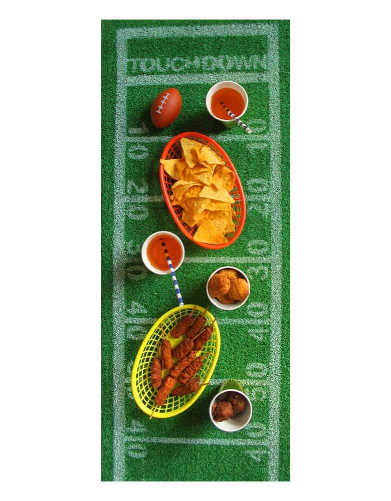 A overhead shot of a football party with Momopartys's 5ft x 16 inches Football Turf Table Runner by Talking Tables. On the table runner there are multiple football game snacks including chicken wings, chips and drinks. This grass runner features yard line markings and touchdown zones just like a football field. Perfect as a football party decoration for the Super Bowl party, game day or a kid's football-themed birthday party.