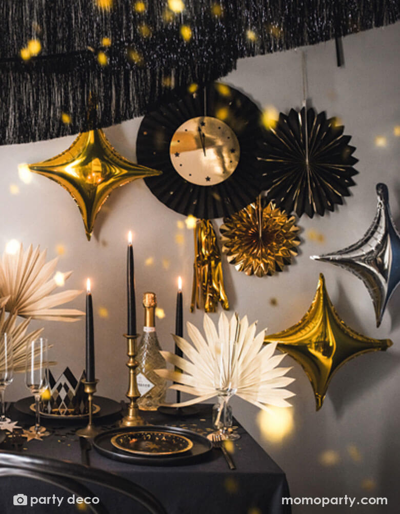 A glamorous black and gold New Year party set up featuring Momopartys's New Year Countdown Party Fans, starpoint shaped balloons and black fringe garlands hung on the wall. In the front is a party table adorned with beautiful black and gold candles, confetti, new year party tableware and floral arrangement, making this a perfect inspiration for a new year eve countdown party.