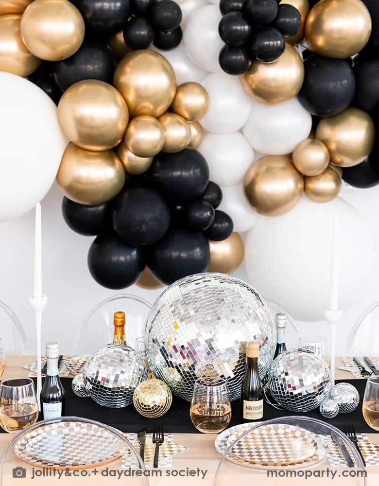 A gorgeous New-Year's-Eve-Party-Table set up featuring a balloon garland of black, gold and silver balloons, table set with Momopartys's silver and gold checkered tableware including plates, napkins and cups, in the middle there are some disco ball decorations in different sizes as the centerpiece, making this a chic and glimmer look for New Year's Eve countdown party. 