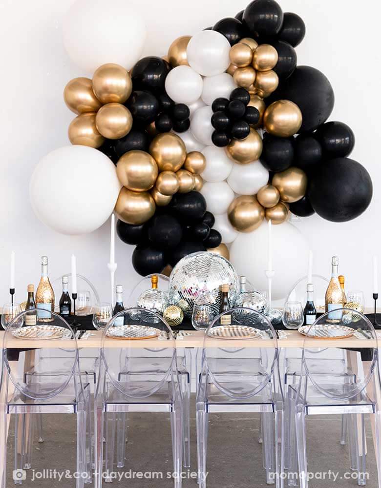 A gorgeous New-Year's-Eve-Party-Table set up featuring a balloon garland of black, gold and silver balloons, table set with Momopartys's silver and gold checkered tableware including plates, napkins and cups, in the middle there are some disco ball decorations in different sizes as the centerpiece, making this a chic and glimmer look for New Year's Eve countdown party. 