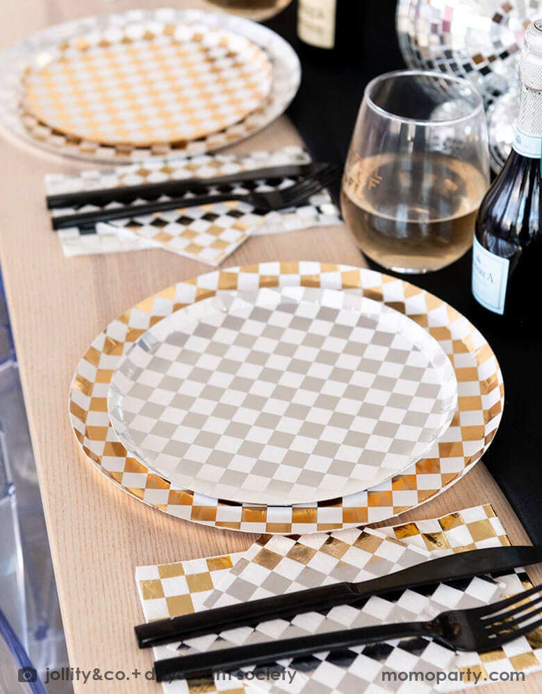 A glamorous and dazzling party table featuring Momopartys's check it! silver and gold checkered tableware collection by Jollity & Co. including 10" dinner plates, 8" dessert plates, 6.5" large napkins, and black cutlery. In the middle of the table are a bunch of disco balls in different sizes with silver confetti scattered, what a festive party table! A perfect inspo for New Year's Eve celebration or a disco themed party!