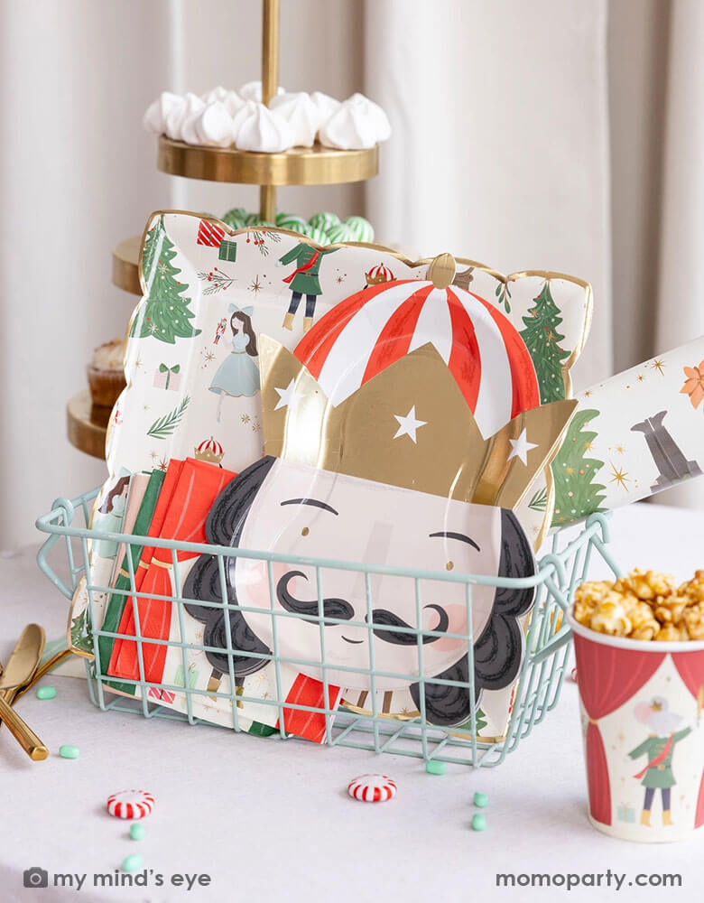 A festive Nutcracker themed Holiday party table featuring an occasion bin filled with  Momopartys's Nutcracker party supplies including the Nutcracker pattern plate, Nutcracker head shaped plate, Nutcracker napkins, Nutcracker paper table runner roll, Nutcracker party cups which are filled with popcorn. On the table there are some Holiday treats including  green and red candies, peppermints, meringues and cupcakes making this a festive Christmas party table for kids and guests of all ages!