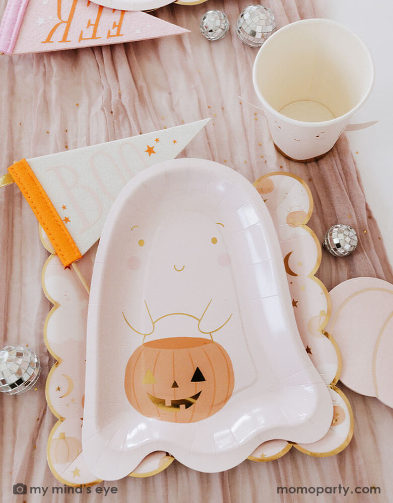 A Halloween party table filled with pink Halloween ghost themed party supplies from Momopartys including the pink ghost shaped paper plates with a jack-o-lantern, the 10" x 10" pink ghost scattered scalloped dinner plates, ghost party cup and Boo and EEK felt party pennants. With mini disco ball decorations, it makes a great inspo for a kid's friendly pink Halloween party this spooky season!