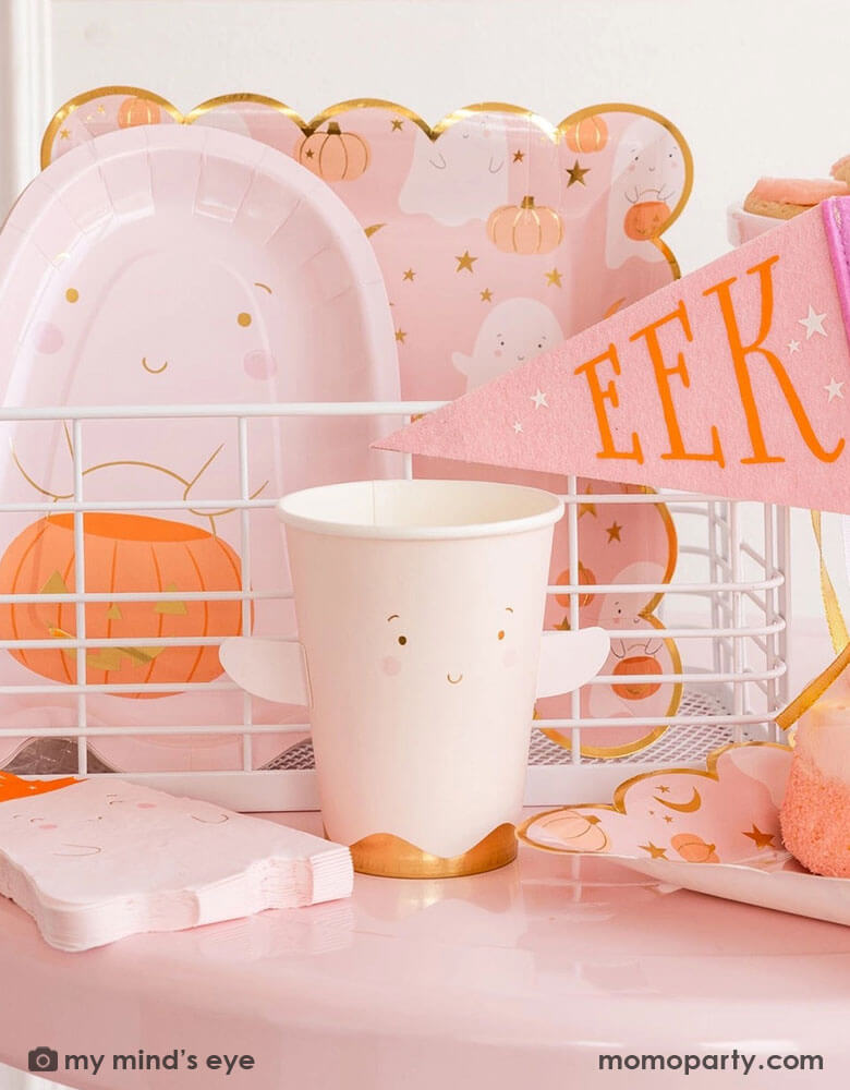 A pink Halloween party table featuring multiple adorable ghost themed party supplies from Momopartys including the 12 oz pink ghost shaped party cup with adhesive arm attachment, the ghost scattered scallop dinner plates, some ghost shaped napkins, a pink ghost shaped die cut plate with a smiley ghost holding a jack-o-lantern for treat or trick. Along with the pink felt party pennant which says "EEEK" on it, it makes a great inspo for putting together a not-so-spooky kid's Halloween party.