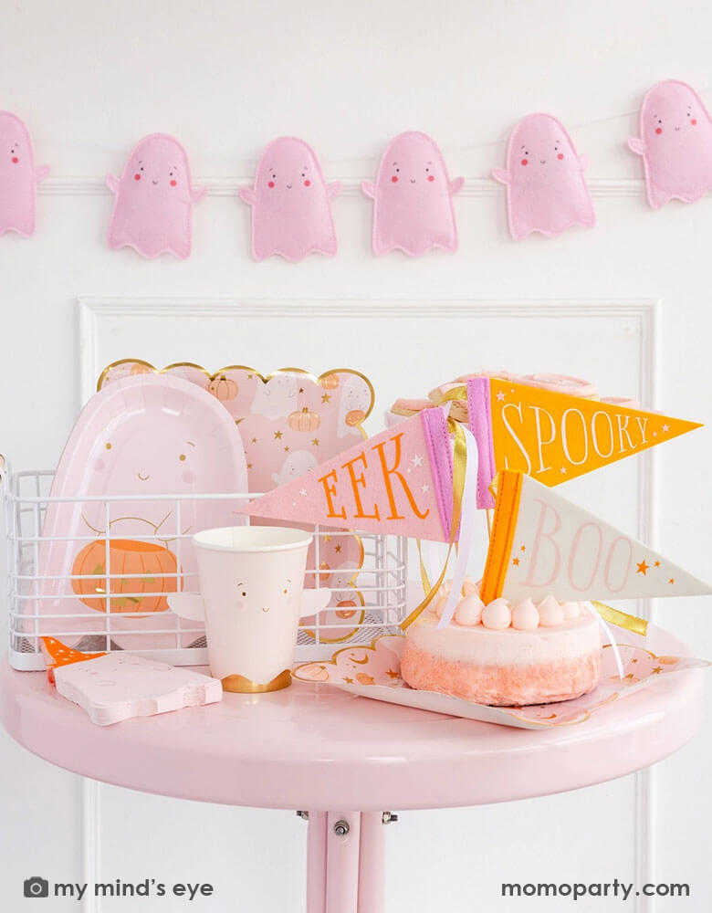 A pink Halloween party table featuring multiple adorable ghost themed party supplies from Momopartys including the 12 oz ghost shaped party cup, the ghost scattered scallop-edged dinner plates, ghost shaped napkins, a pink ghost shaped die cut plate with the ghost holding a jack-o-lantern. Along with a pink cake topped with felt pennants that have "SPOOKY," "EEK" and "BOO" on them, plus the pink felt ghost banner hung on the wall behind. Perfect for a kid's friendly Halloween bash!
