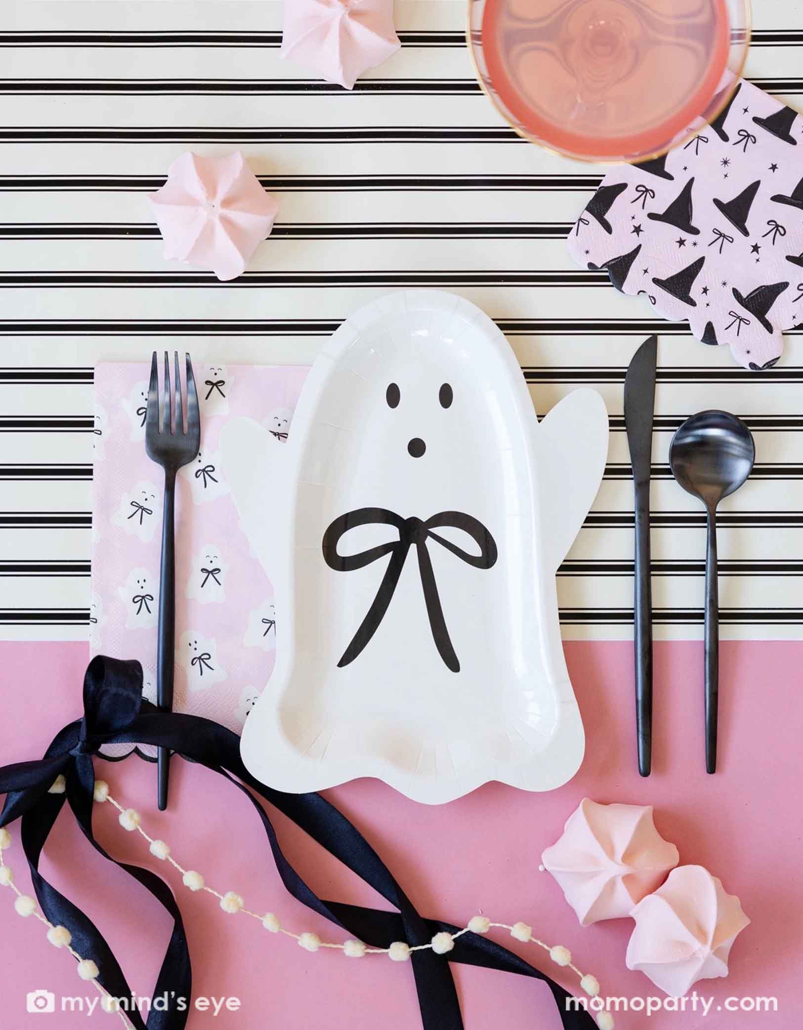 A overhead shot of a pink Halloween themed party tablescape featuring various adorable pink themed Halloween party supplies from Momopartys including the Halloween ghost plate with black bow, the pink napkin with witch hat and black bow pattern on it, black fork, knife, and spoon on a white and cream striped tablerunner with beautiful black bow around and light pink meringue desserts on the table.