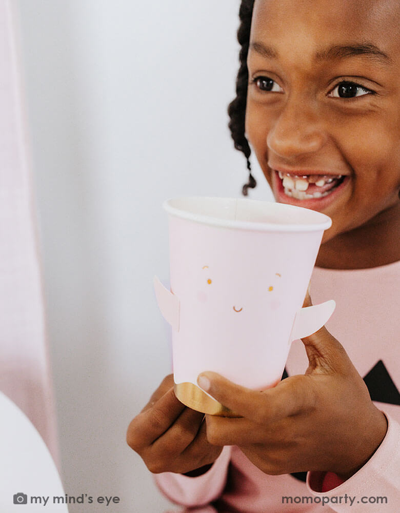 A school age girl holding Momopartys's 12 oz Ghost Paper Party Cup by Occasions By Shakira. An adorable and cute party cup for a kid's friendly pink Halloween celebration in the spooky season.