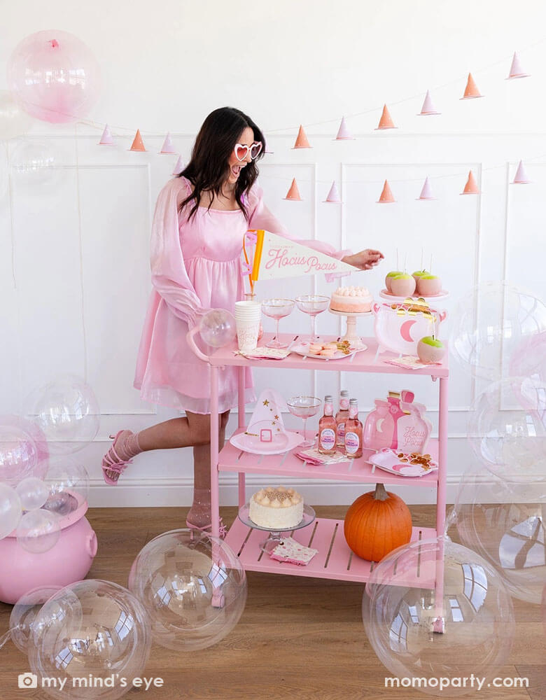 A pink Halloween party with a lady in her pink outfit and pink heart shaped sunglasses. There's a pink cart filled with Momopartys's pink witch party supplies including the Hocus Pocus felt party pennant, party cups, pink cauldron shaped plates, pink witch hat shaped plates and pink potion shaped plates. On the wall hung the 3D pink witch banner. On the floor there's a pink witch's cauldron with clear balloons as if bubbles coming out of it. This gives a great inspo for a fun pink Halloween bash for girls.