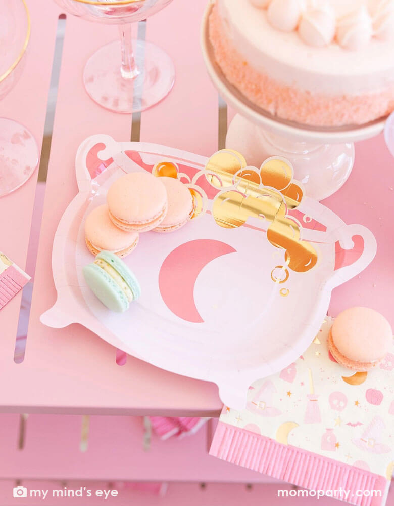 A pink Halloween party table featuring Momopartys's pink witch themed party supplies by My Mind's Eye including the pink cauldron shaped plate and the Hocus Pocus pink fringe napkin. On the table there are also some pastel colored macarons on the plate and the napkin which had a pink buttercream cake and some pink glasses next to them.  This pink table is a perfect inspo for a fun and chic pink Halloween party for girls in the spooky season!
