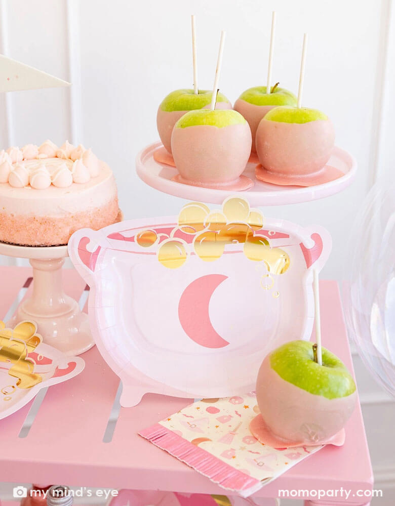 A pink Halloween party table featuring Momopartys's pink witch themed party supplies by My Mind's Eye including the pink cauldron shaped plates, Hocus Pocus pink fringe napkins, and Hocus Pocus felt party pennant, on the table there are also some pink dipped caramel apples and a pink buttercream cake, making this a perfect inspo for a fun and chic pink Halloween party table decoration idea.