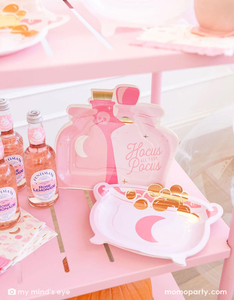 A pink party cart filled with pink Halloween witch themed party supplies from Momopartys including the pink cauldron shaped plates, the pink potion shaped plates, and the Hocus Pocus pink fringe small napkins by My Mind's Eye. Around the tableware there are some rosie pink lemonade bottles and some pink caramel dipped apples, making this a perfect inspo for a chic and stylish Halloween celebration for girls of all ages.